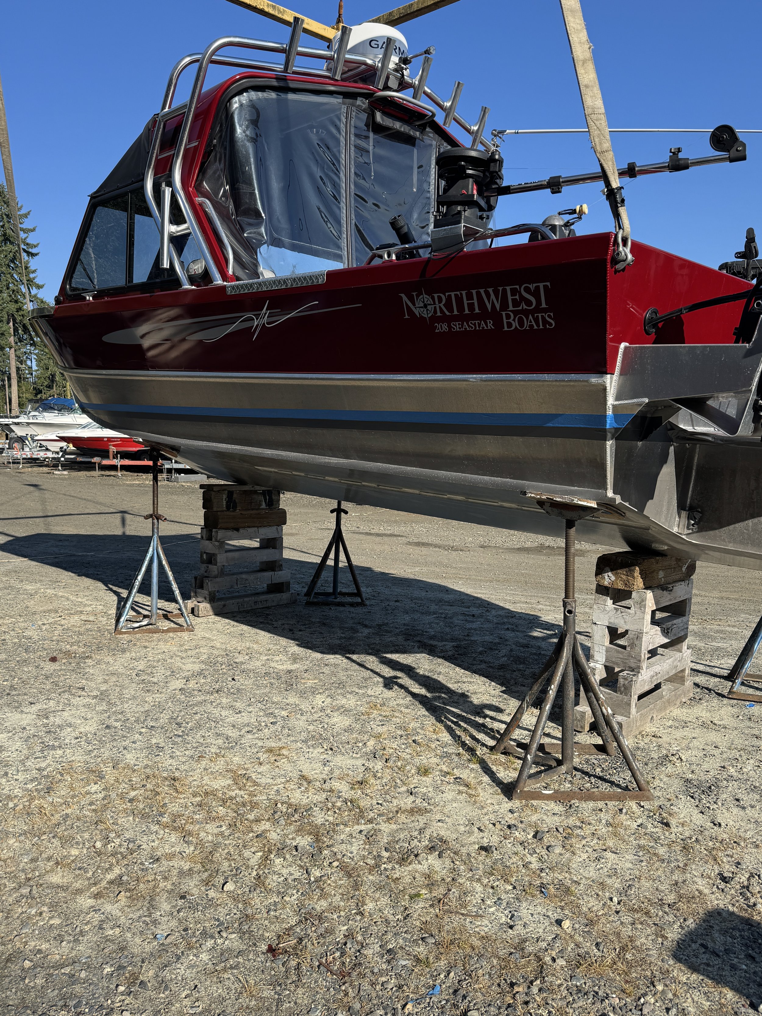 Sand Blasted A red and black boat on land supported by wooden pallets and metal stands, under a clear blue sky.