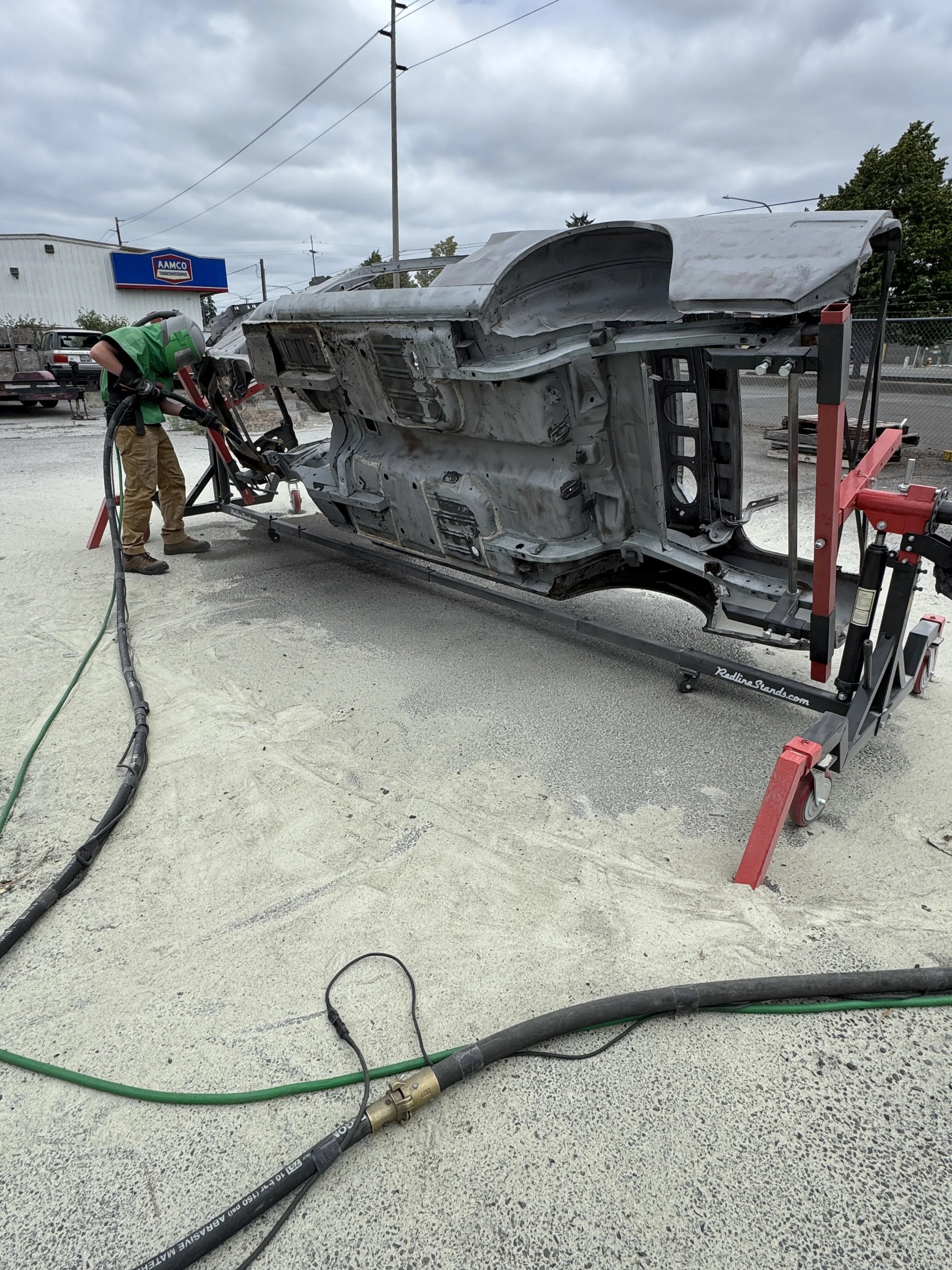 sand blasting a mustang, A worker in green shirt and tan pants is welding or grinding the underside of a car frame, which is lifted on a hydraulic stand outside a workshop. The scene includes tools, hoses, and a cloudy sky.