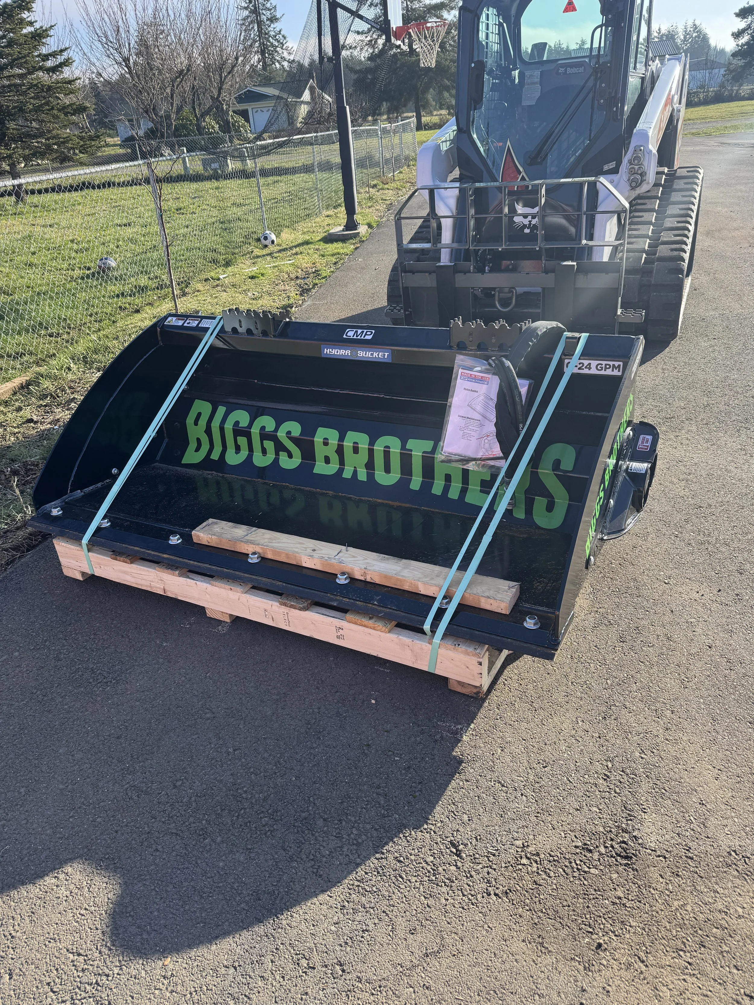 A skid-steer loader attached to a black Biggs Brothers snow plow with green lettering. The plow is on a wooden pallet on a paved driveway. The background includes a chain-link fence, grass, trees, and houses.