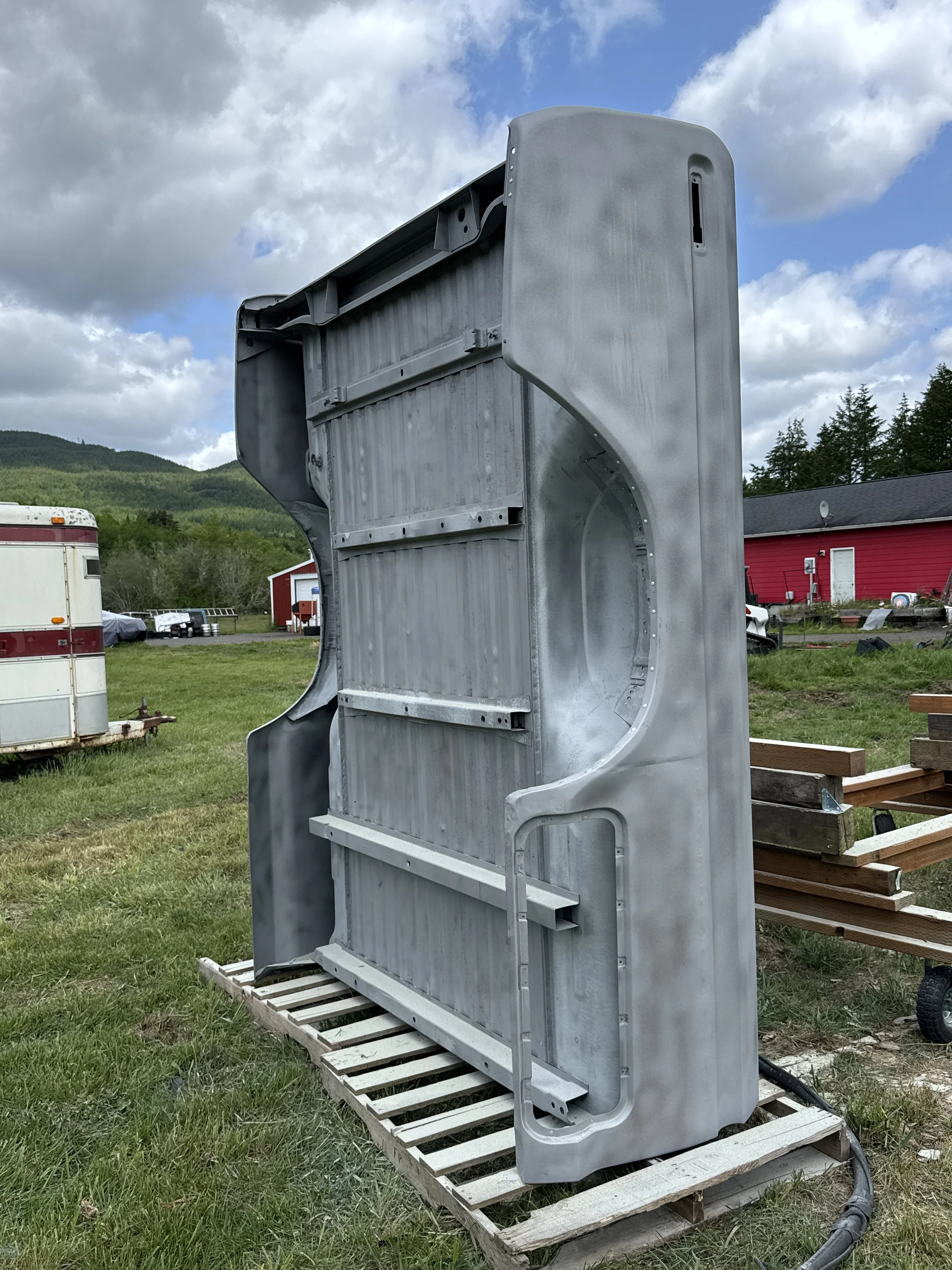 sand blasted a ford truck bed, standing vertically outdoors on a wooden pallet, with a green grassy area, trailers, a red building, and a cloudy sky in the background.