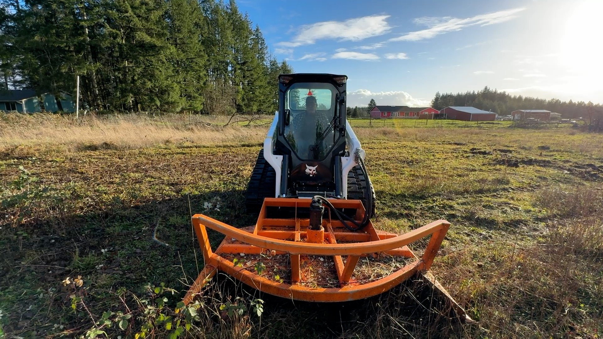 A Bobcat t62 track loader  with a Davco 72" brush hog mowing a field, with trees and farm buildings in the background under a partly cloudy sky.