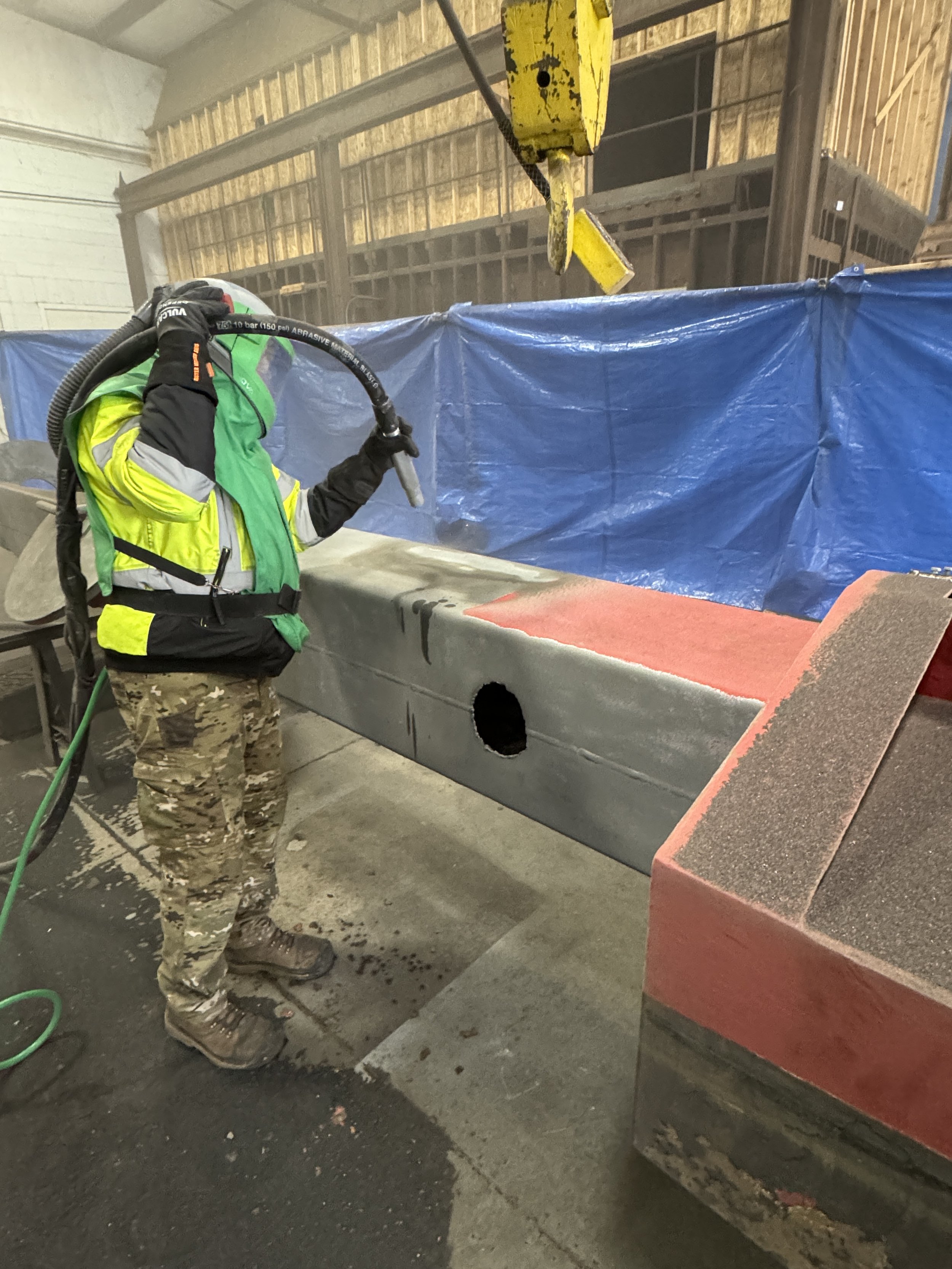 Worker sandblasting a large concrete pipe in an industrial workshop, wearing a safety helmet and reflective clothing.