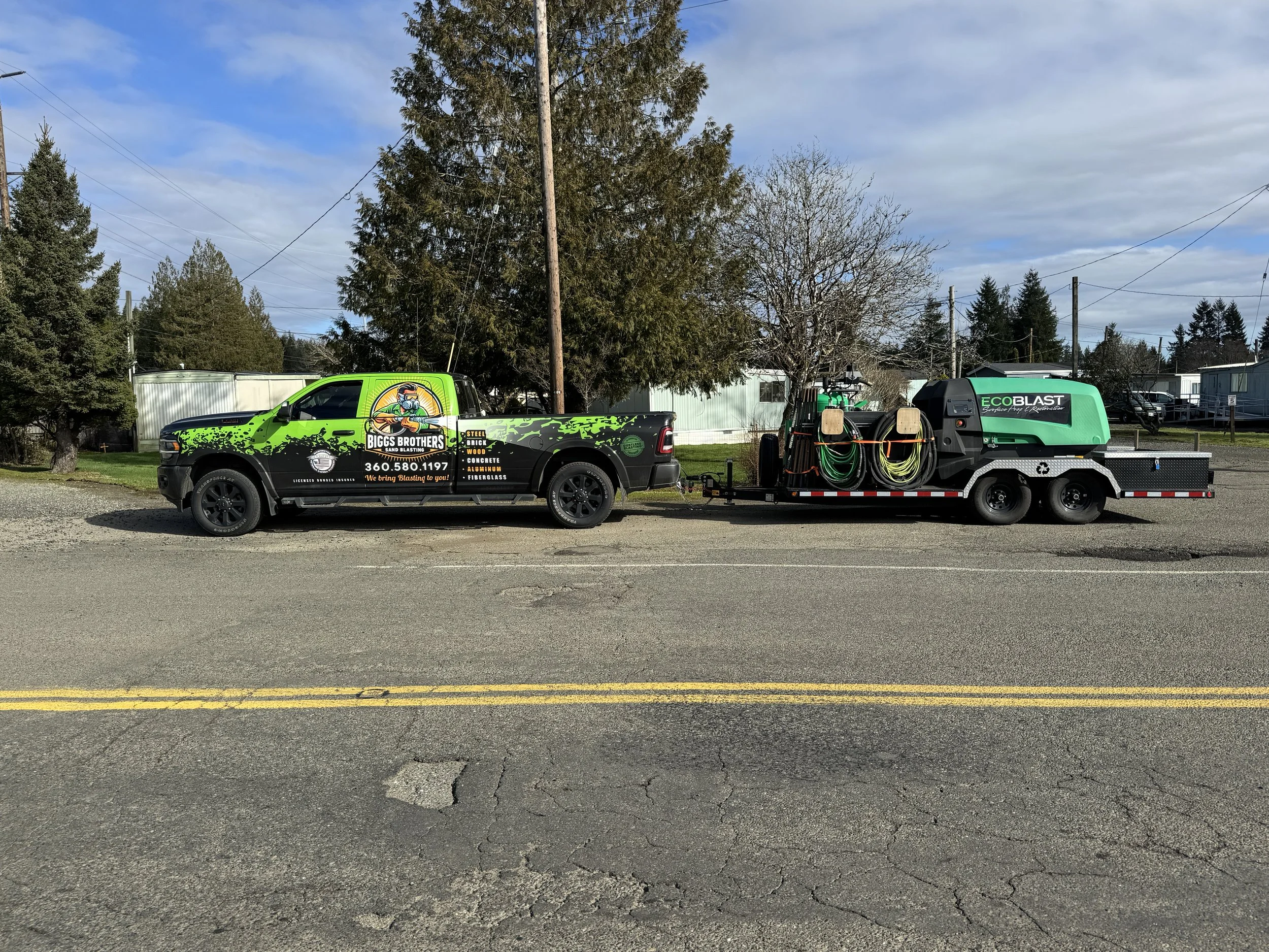 A green and black Biggs Brothers truck attached to a green sand blasting trailer parked on a street. The trailer has hoses and cleaning equipment. Background includes trees, a utility pole, and residential buildings.