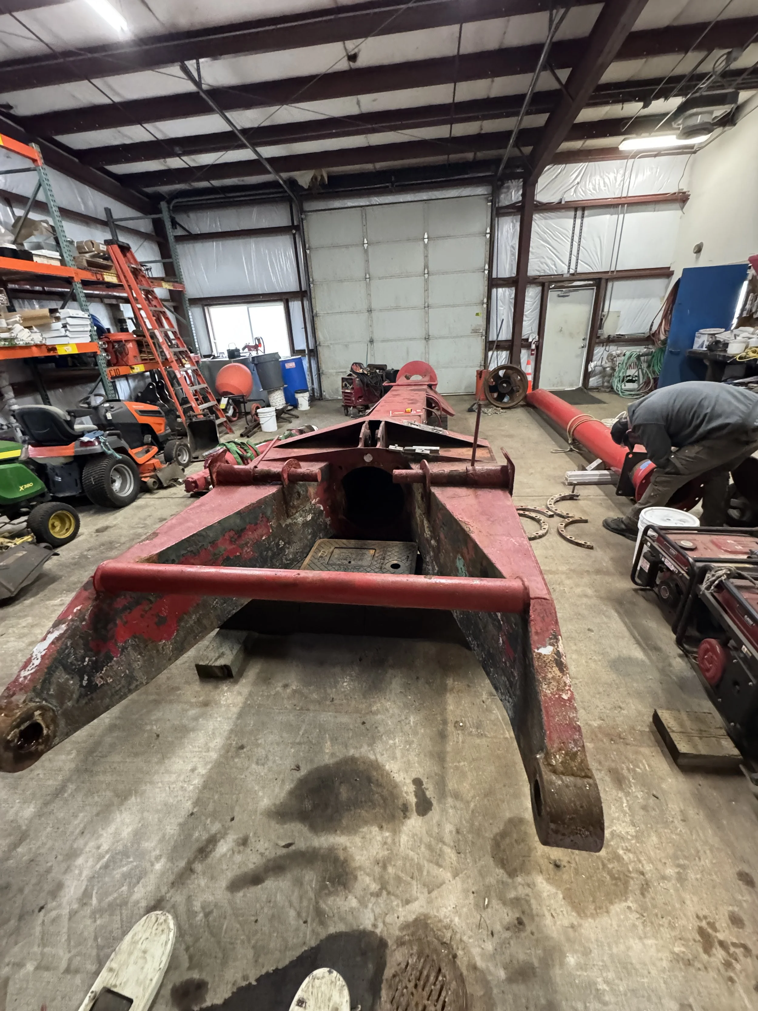 To be sand blasted, The image shows an indoor workspace with various tools and equipment, including a ladder, a lawnmower, and a person working on a large, red, rusted piece of machinery or equipment, possibly a boat or heavy-duty vehicle chassis.
