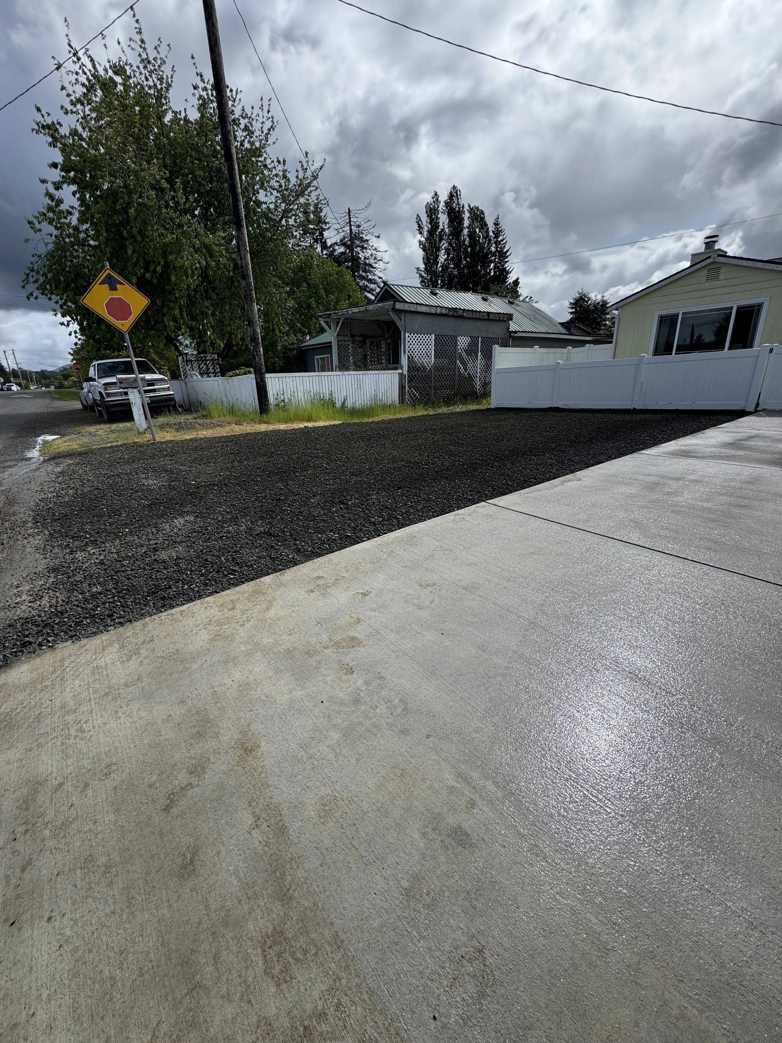 Residential street view showing a driveway with concrete and gravel sections, a stop sign, parked pickup truck, and houses under cloudy sky.