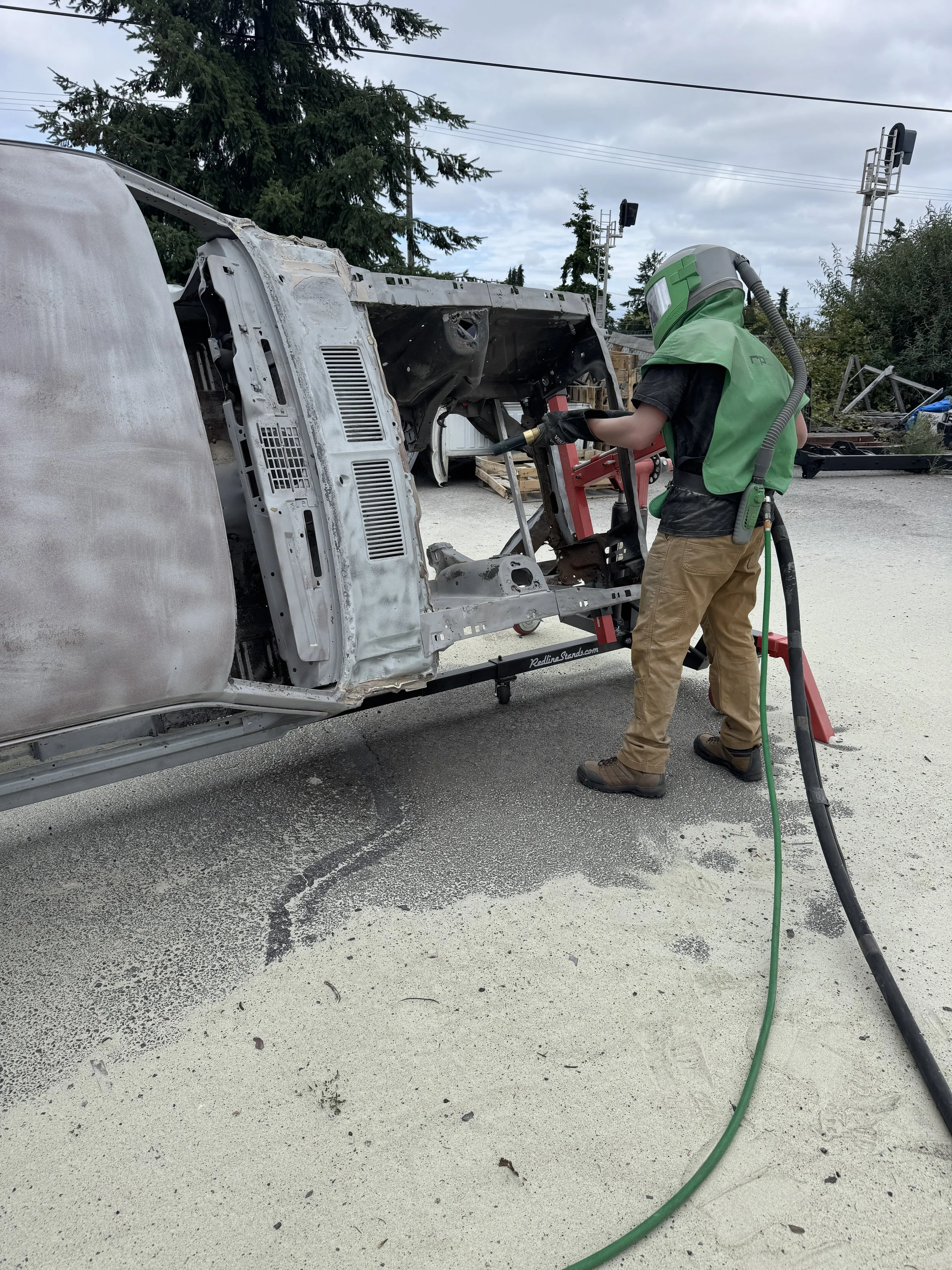 sand blasting a mustang, A person wearing a protective helmet, green vest, and brown pants is sanding the frame of a car body on a portable workbench outdoors, with trees and a cloudy sky in the background.