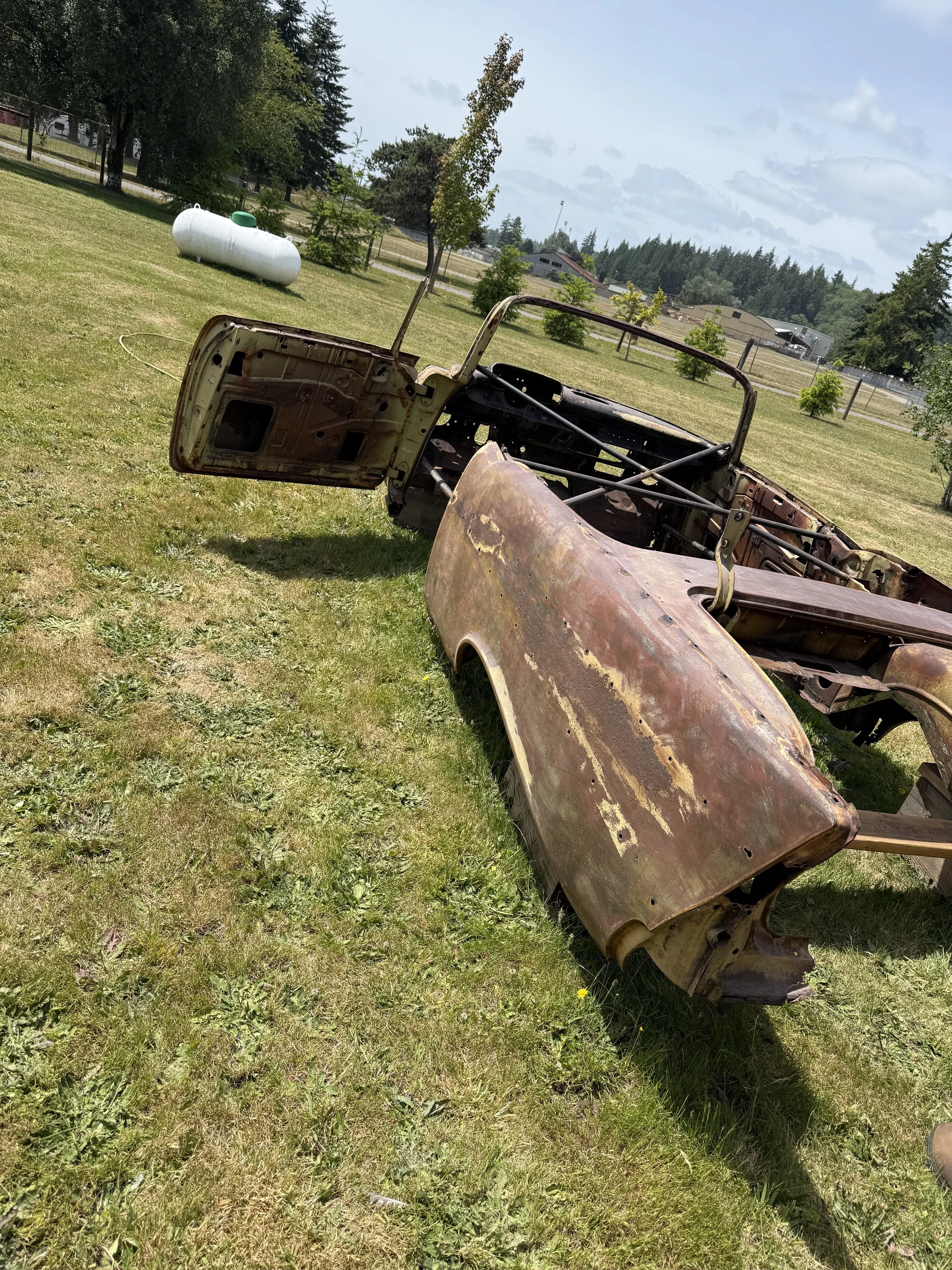 sand blasted a 57 chevy, Rusty, abandoned car frame lying on the grass in a park-like setting with trees and a propane tank in the background.