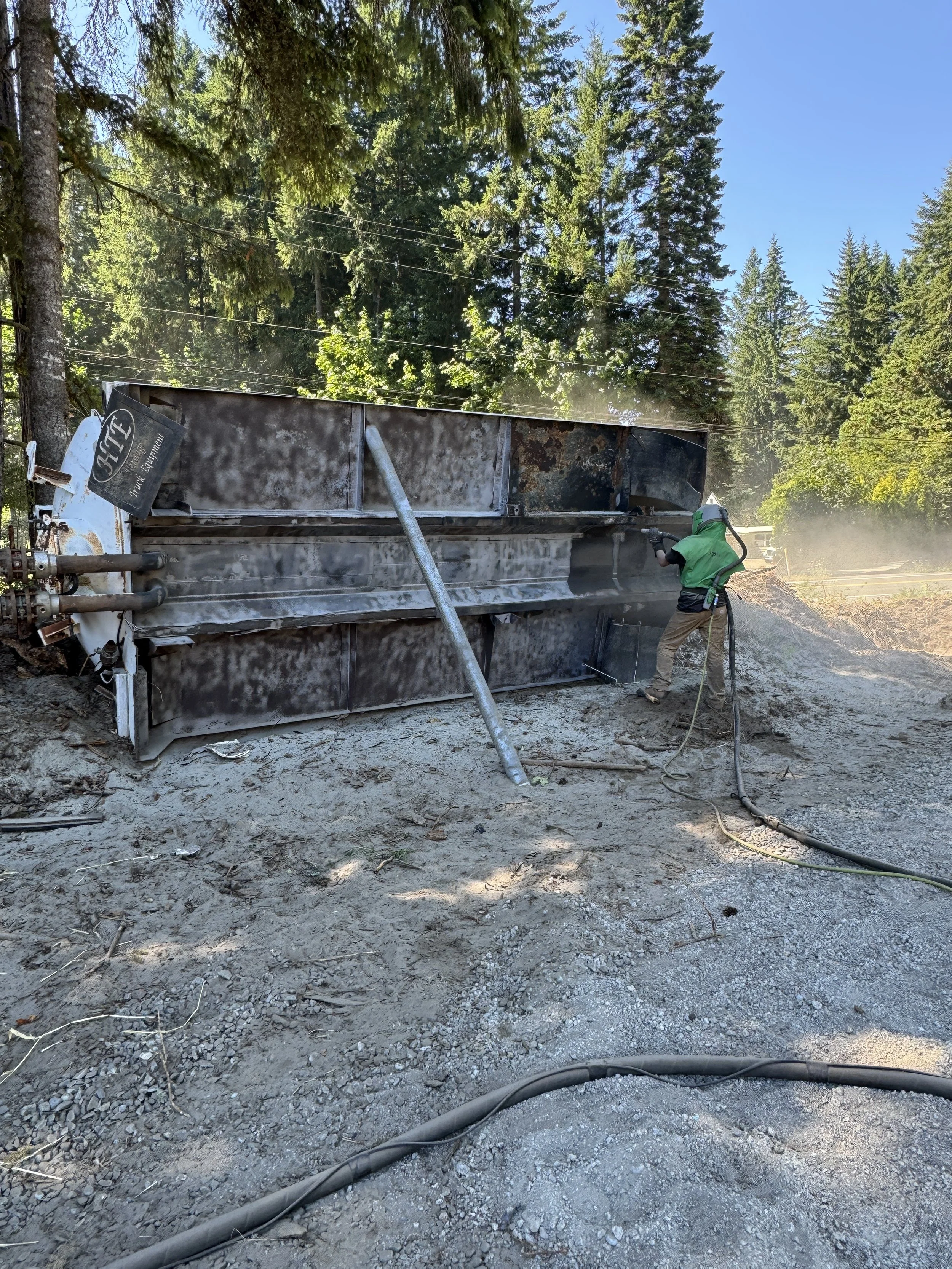 sand blasted tank bottom, A worker in safety gear using a pneumatic tool on a large,  in a forested area with trees and a dirt ground.