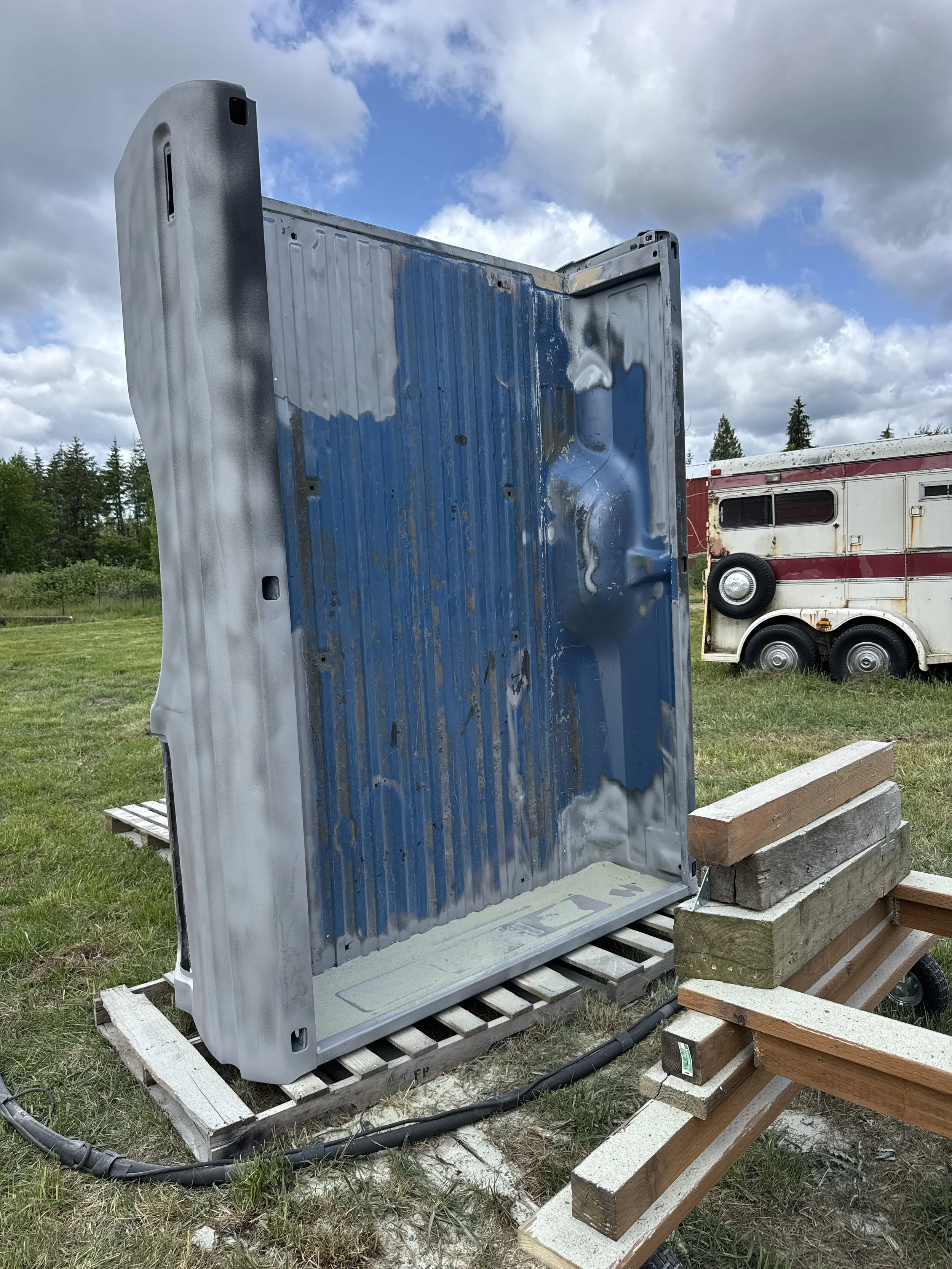 sand blasted a ford truck bed, stands upright on a wooden pallet outdoors. The background features a grassy field, trees, a cloudy sky, and a white and red RV trailer.