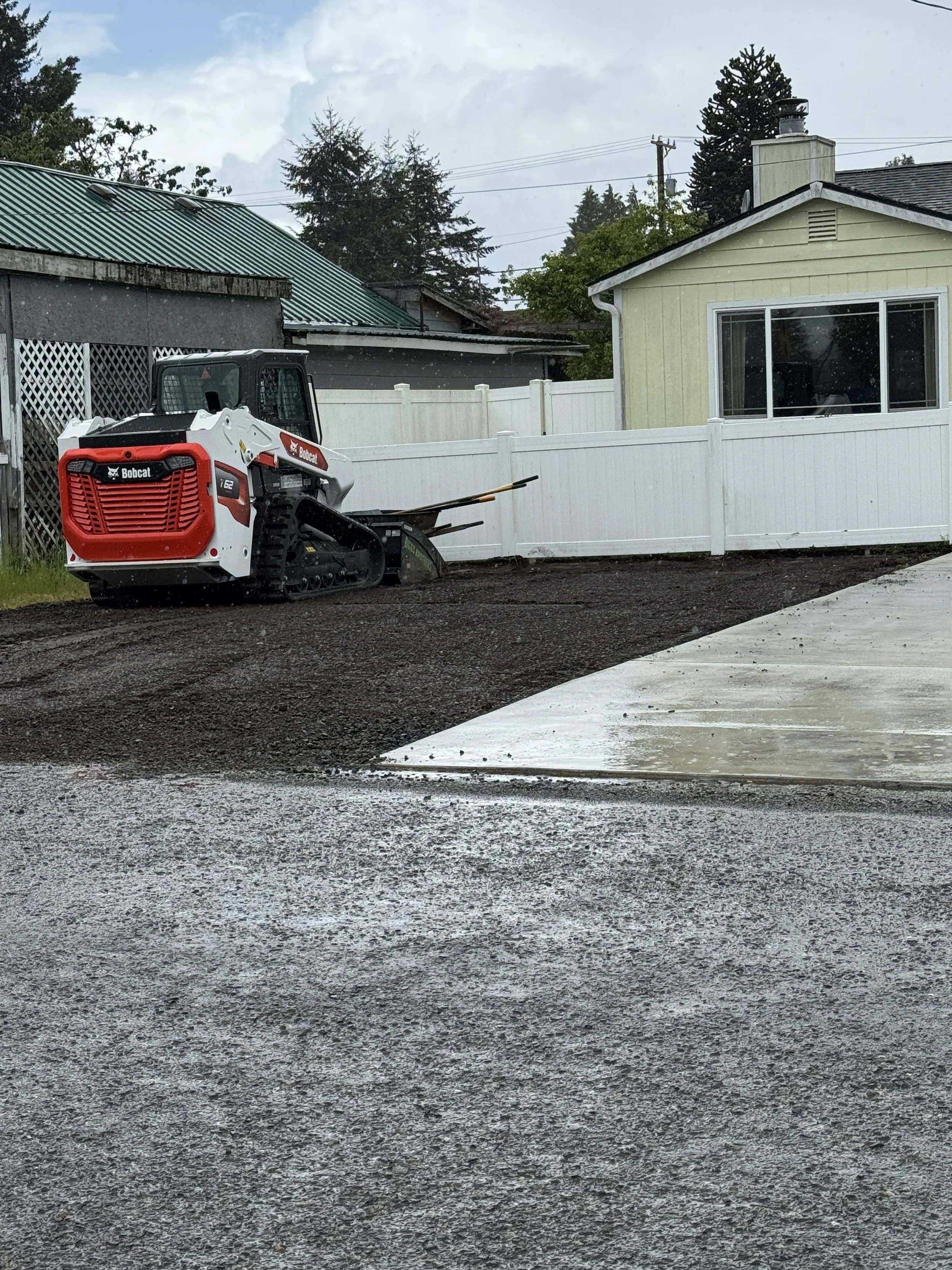 A Bobcat t62 track loader  is grading and preparing the ground for a new driveway behind a yellow house with a white fence, on a rainy day with overcast skies.