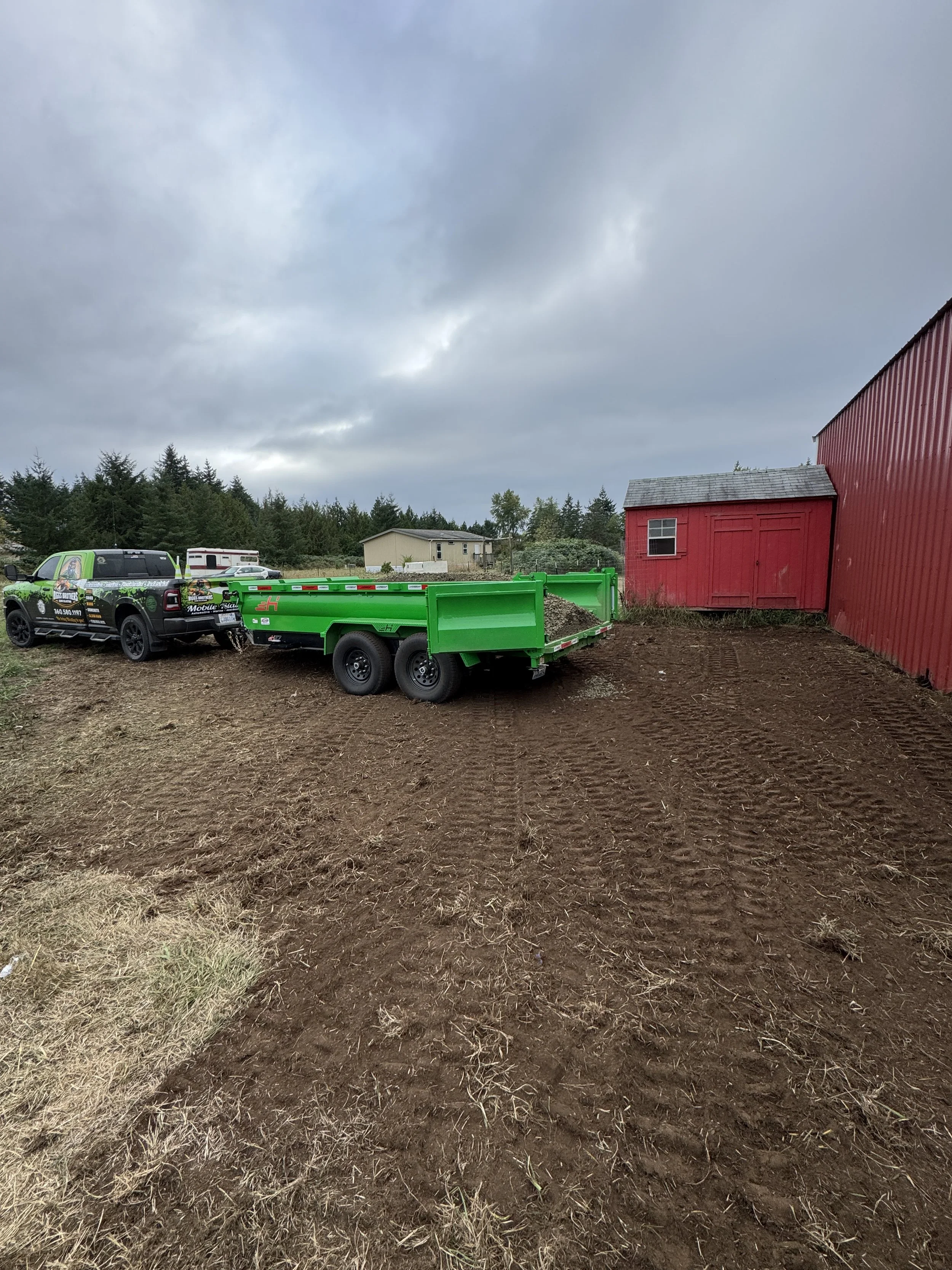 A green trailer attached to a black pickup truck dumping gravel with a colorful logo parked on dirt ground near a red barn. Overcast sky above.