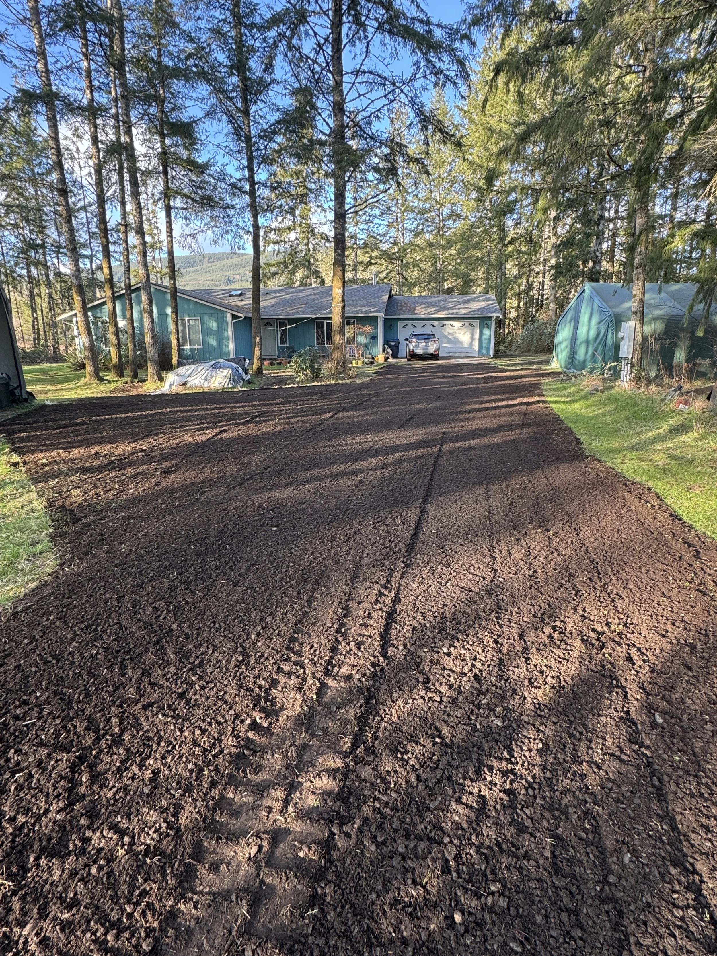 A freshly graded dirt driveway leading to a blue house with white trim, surrounded by tall trees, with a garage door, car, and a green greenhouse on the right.