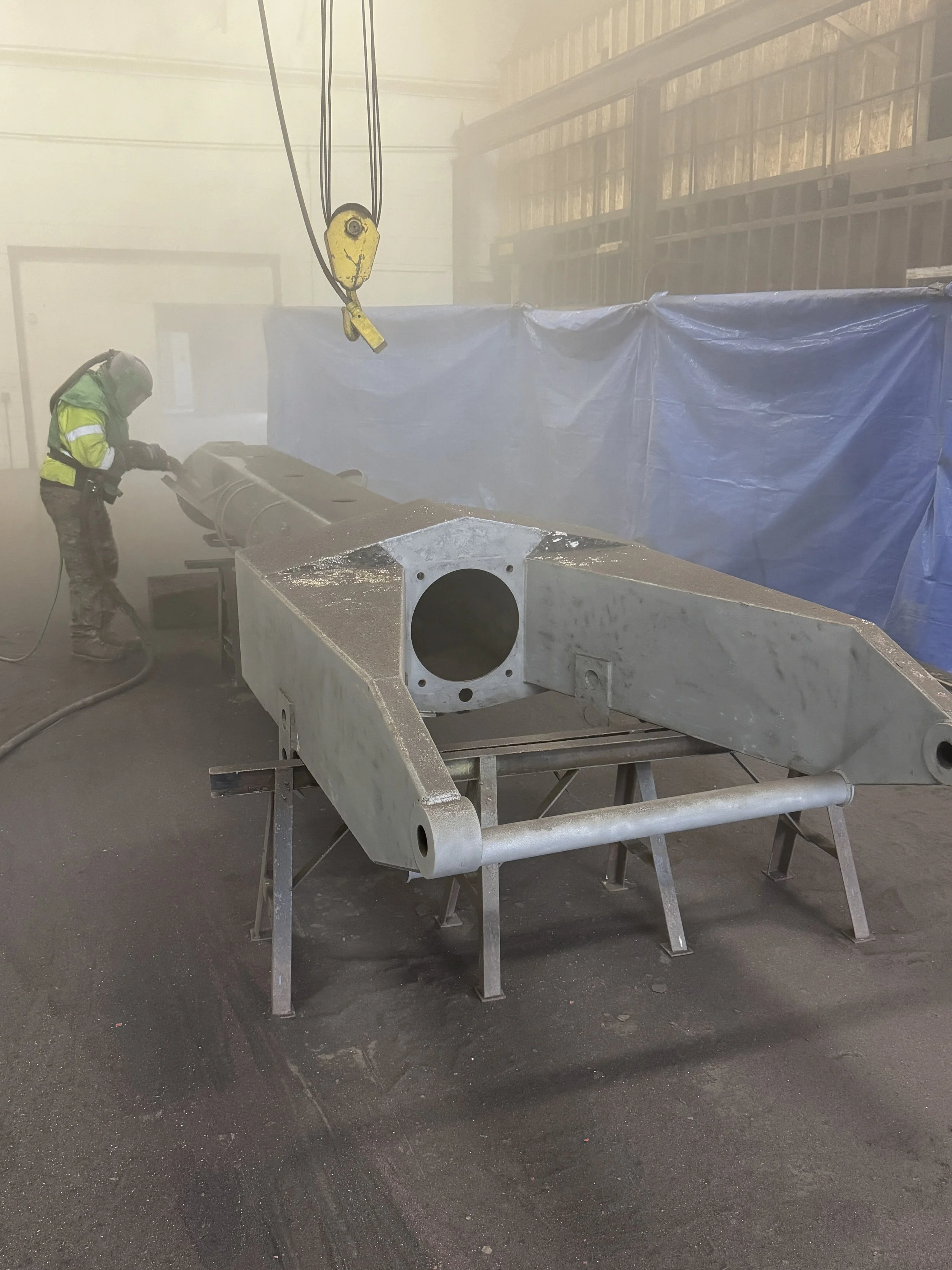 sand blasting, A worker in safety gear cleaning or working on a large metal component inside a workshop, with a blue tarp and industrial equipment around.