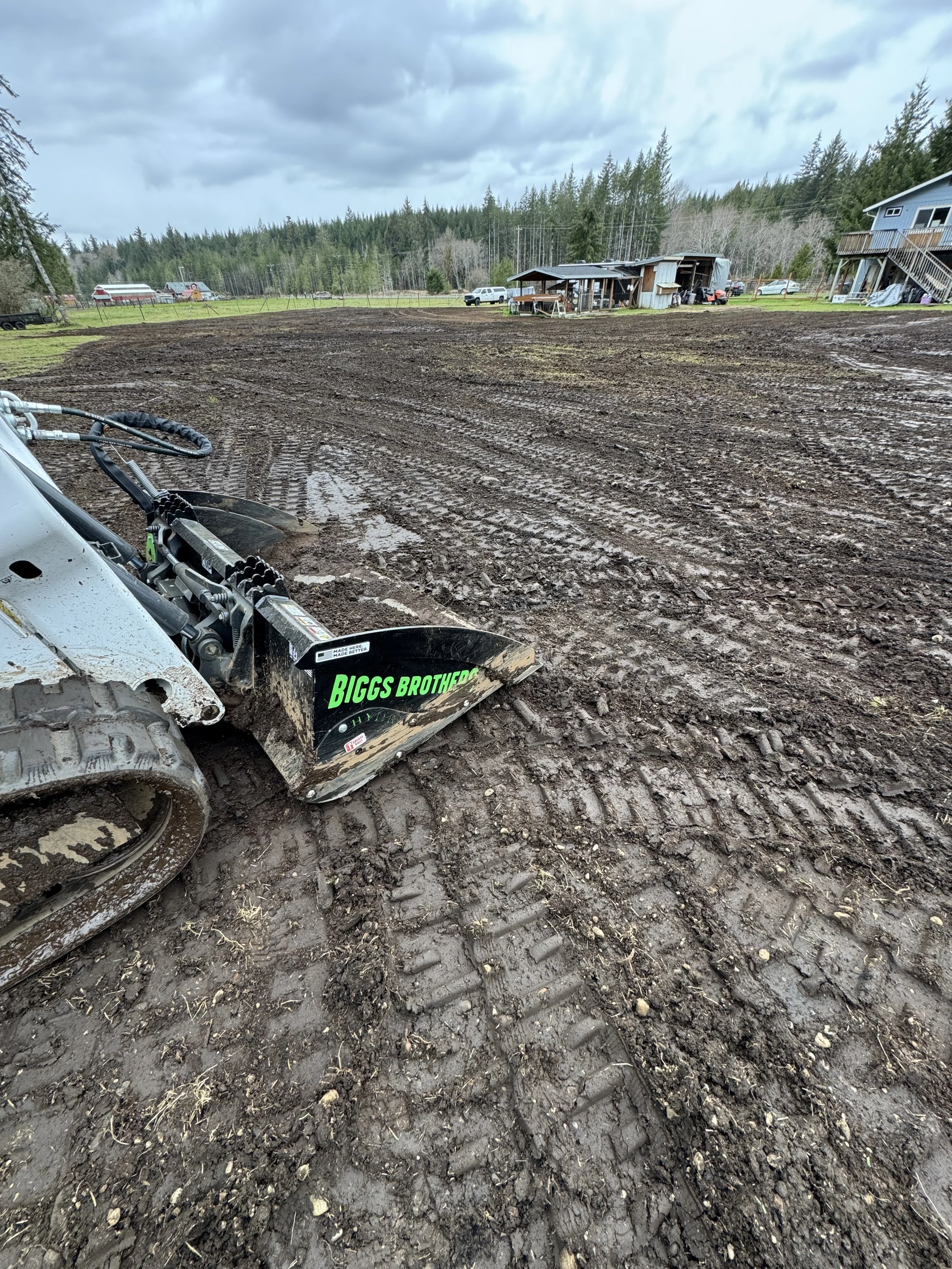 Property cleared for grass, A dirt field with tire tracks from a construction or farming vehicle. In the foreground, there's a piece of machinery labeled "Biggs Brothers." In the background, there are houses and trees under cloudy skies.