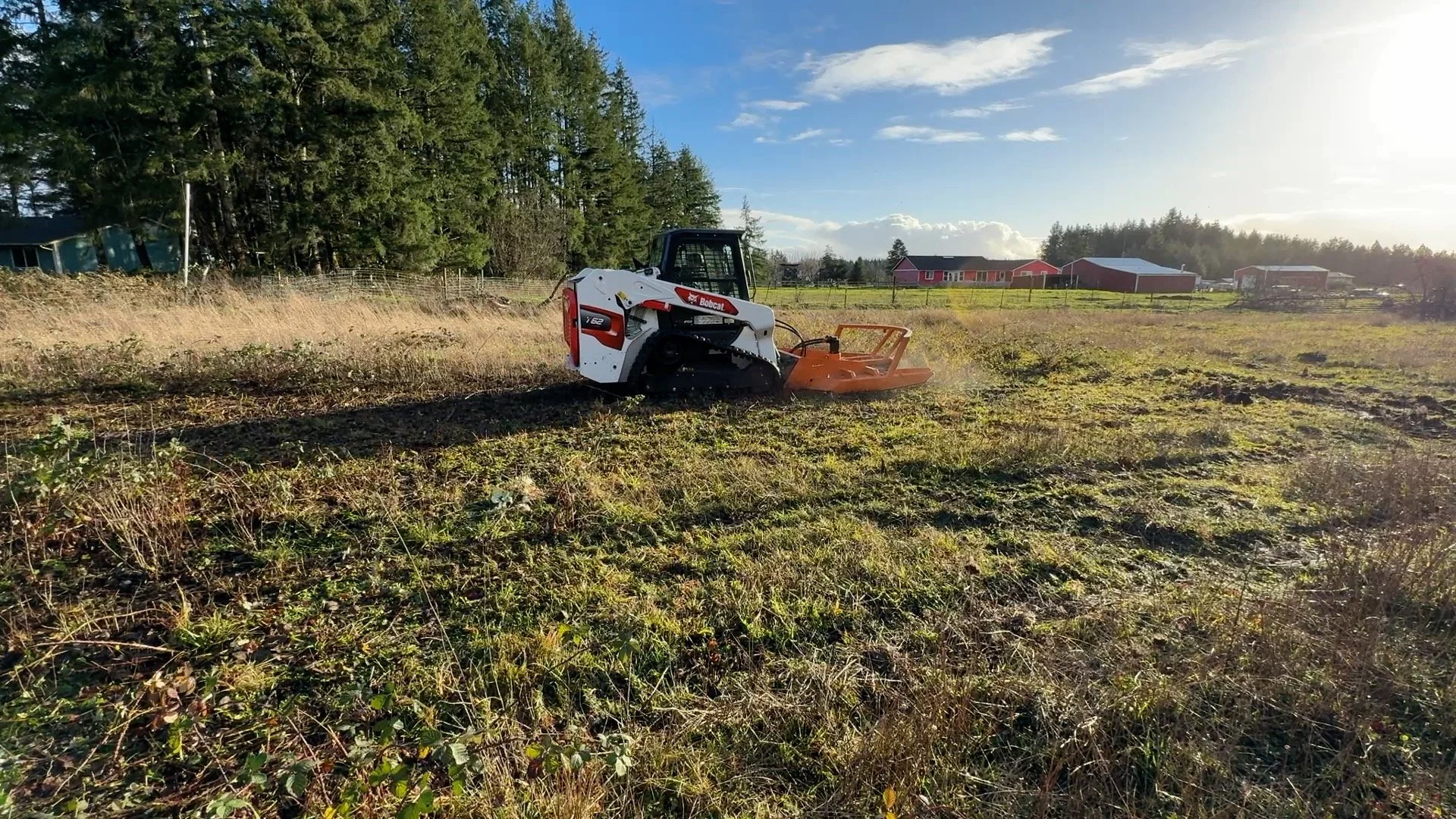A Bobcat t62 track loader  with a Davco 72" brush hogworking on a grassy field with rural homes and a tree line in the background, under a partly cloudy sky.
