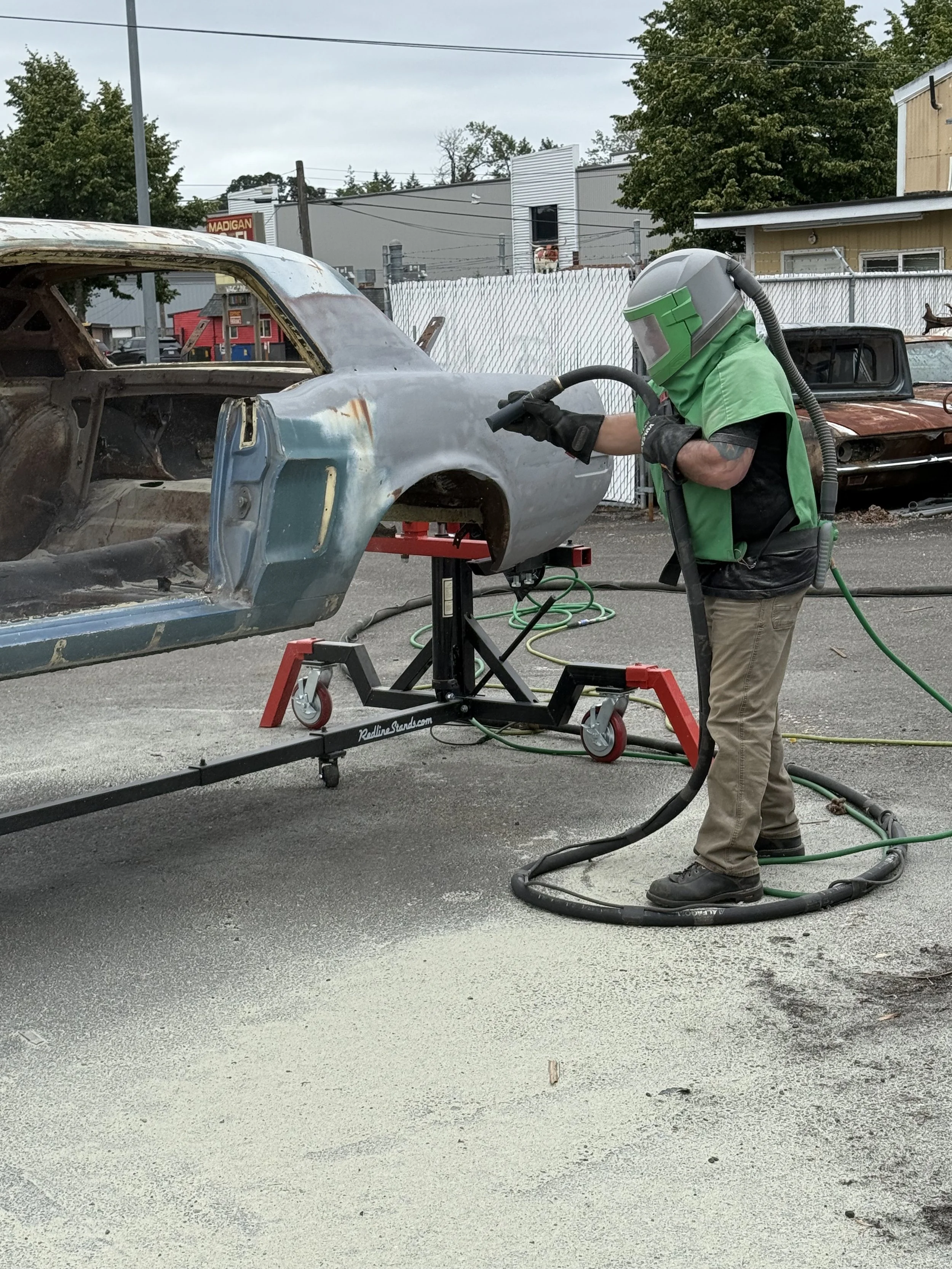 sand blasting a mustang, Person in protective gear grinding the body of a vintage car in an outdoor area with other old vehicles in the background.