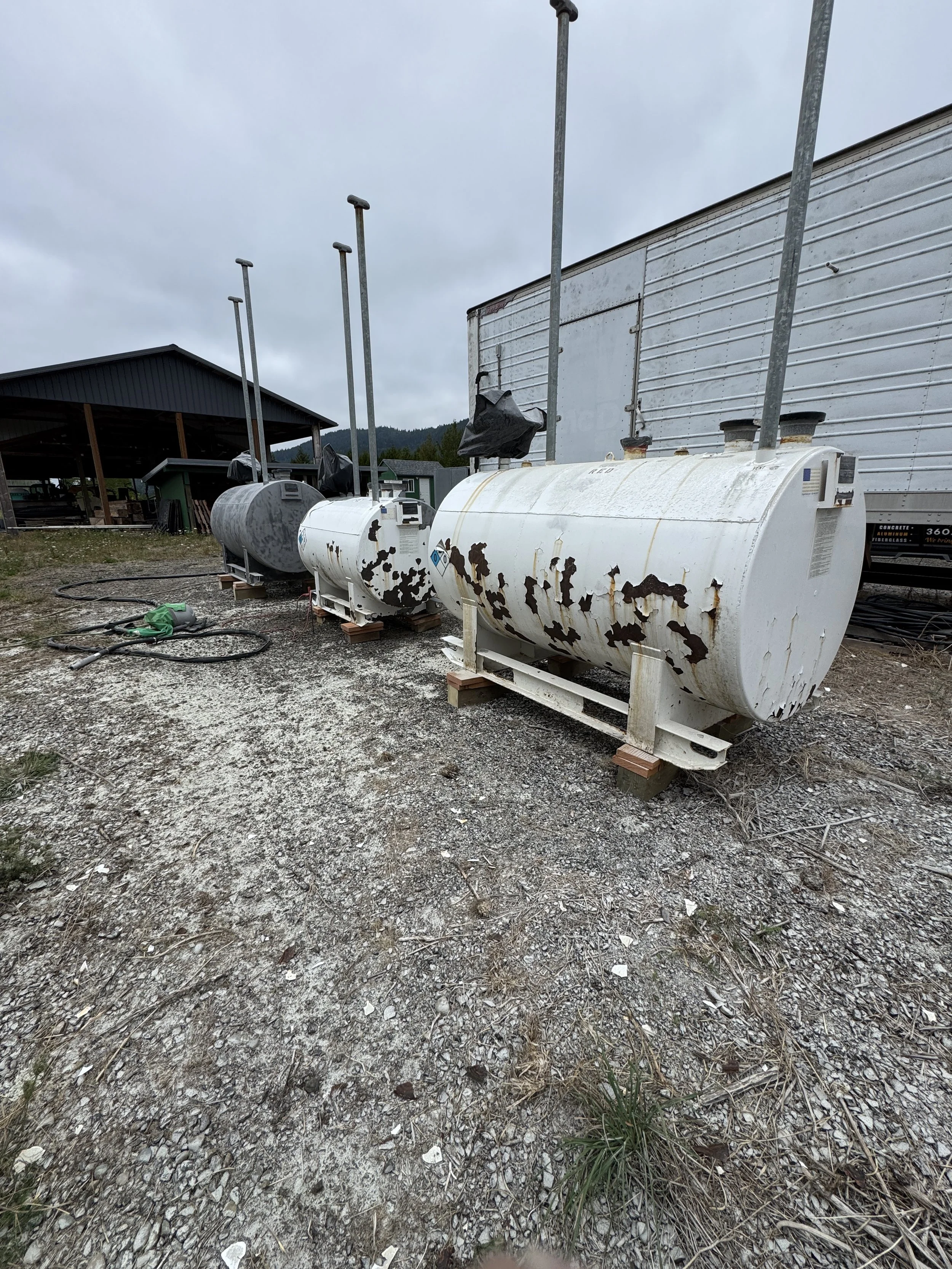 sand blasted Four large, rusted propane tanks aligned outdoors on a gravel surface, with a cloudy sky above and storage structures in the background.