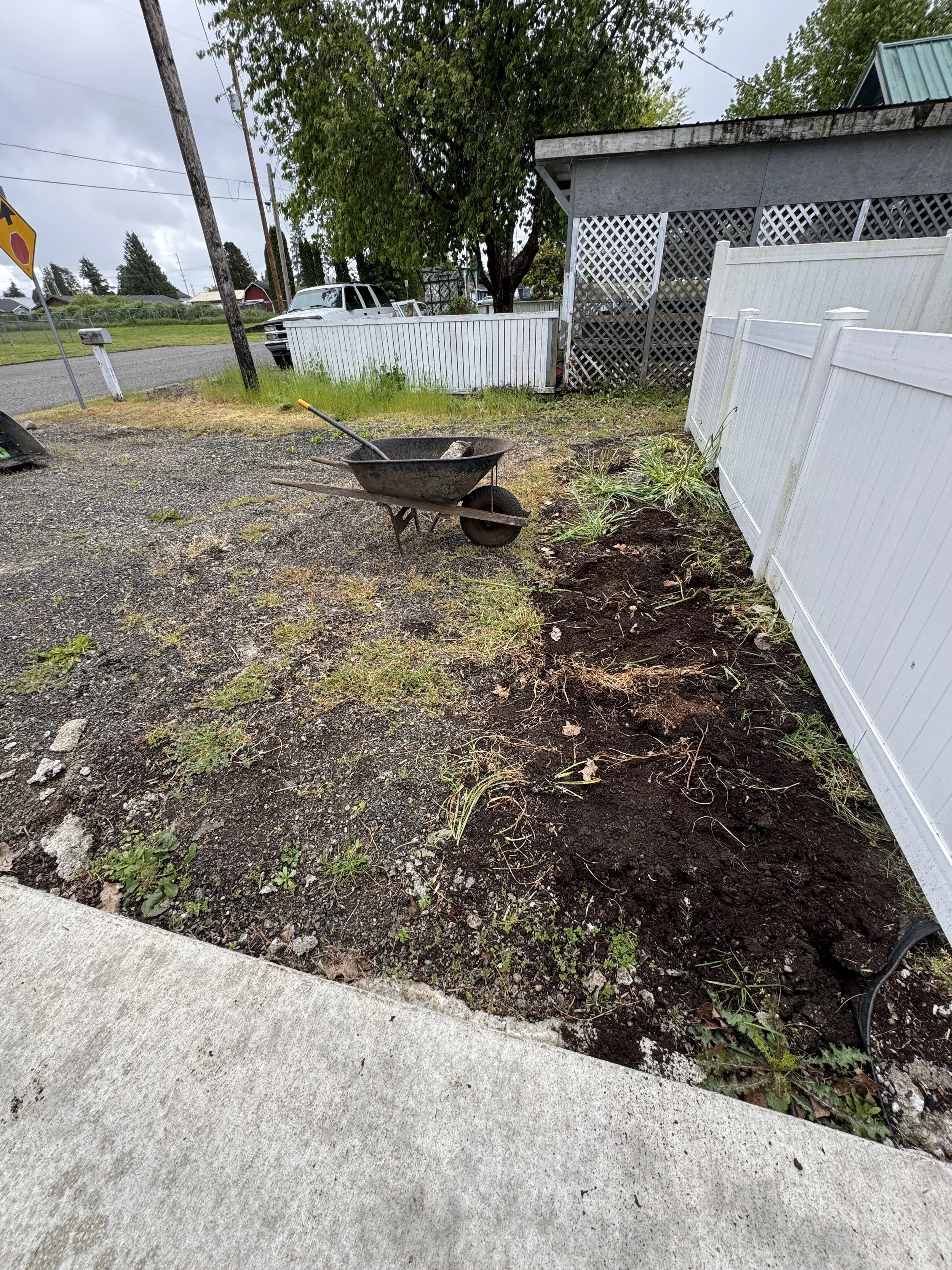 Grasved driveway project, A Bobcat t62 track loader ,being prepared , bordered by a white fence, with a wheelbarrow resting on gravel nearby, in a suburban yard.
