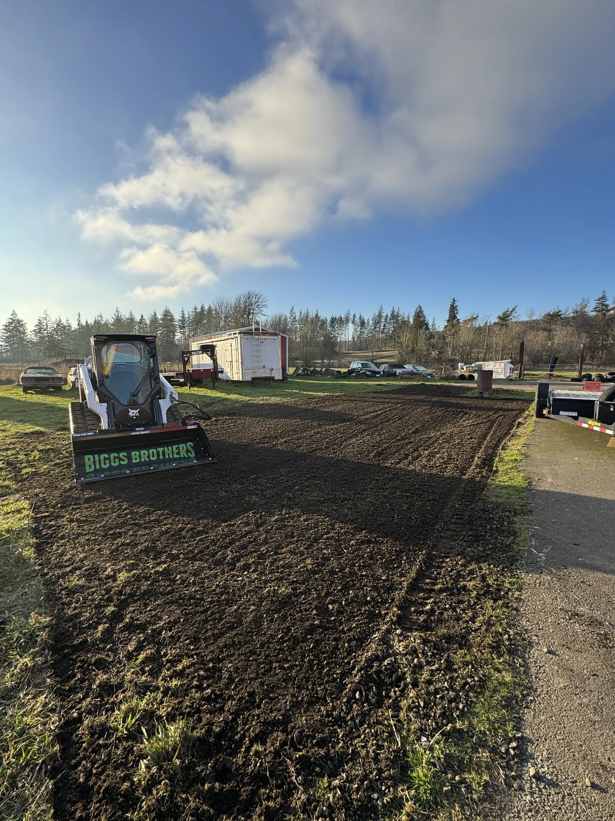 A leveled dirt area with a small loader labeled 'Biggs Brothers' on a sunny day with a partly cloudy sky, in a rural setting with trees and parked trailers in the background.