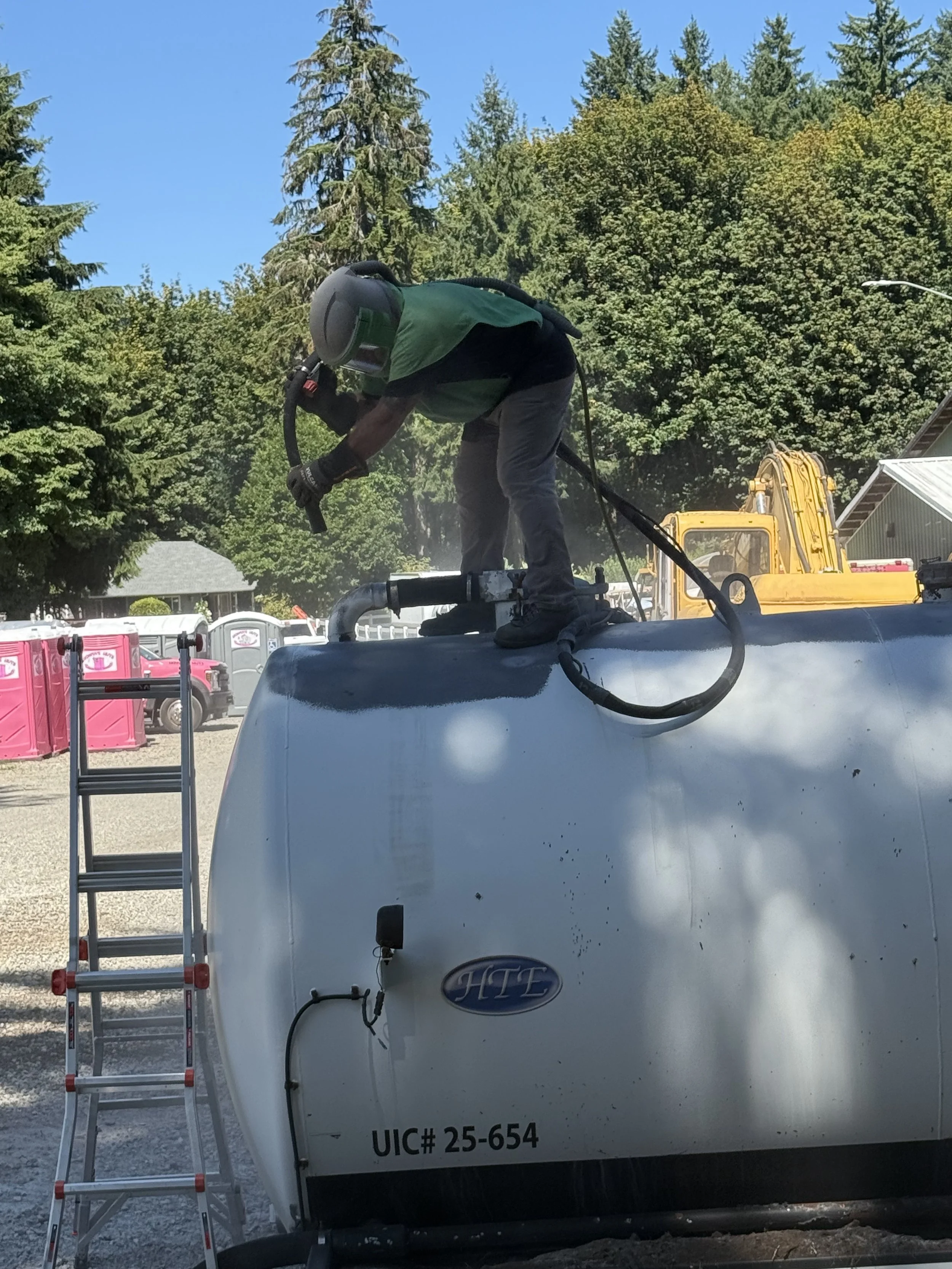 sand blasting a tank, Worker wearing a helmet and green vest cleaning the top of a white tank or trailer with a handheld tool, set outdoors with trees and construction equipment in the background.