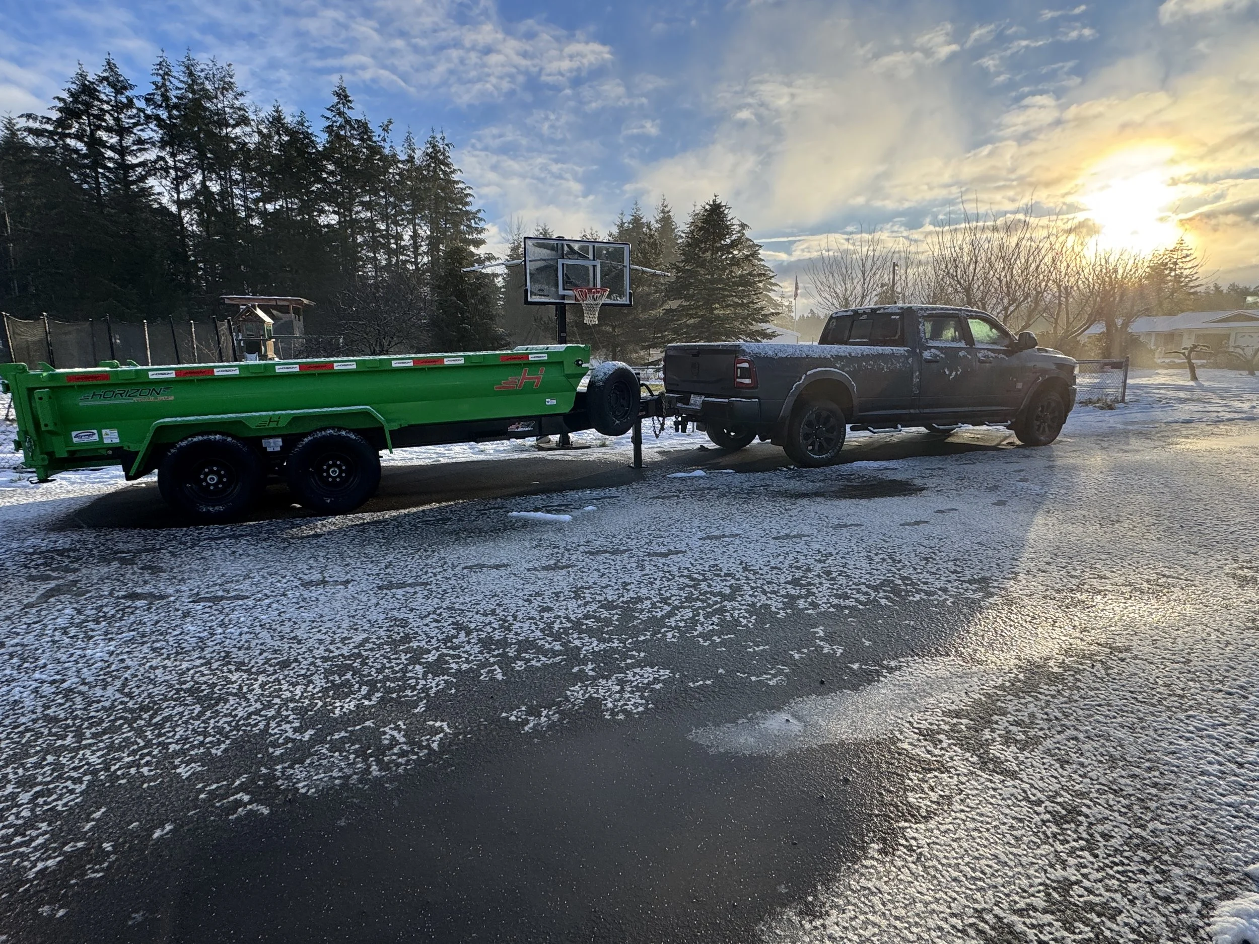 A black pickup truck towing a green trailer parked on an icy driveway with a basketball hoop and a backyard with trees and houses in the background during sunset.
