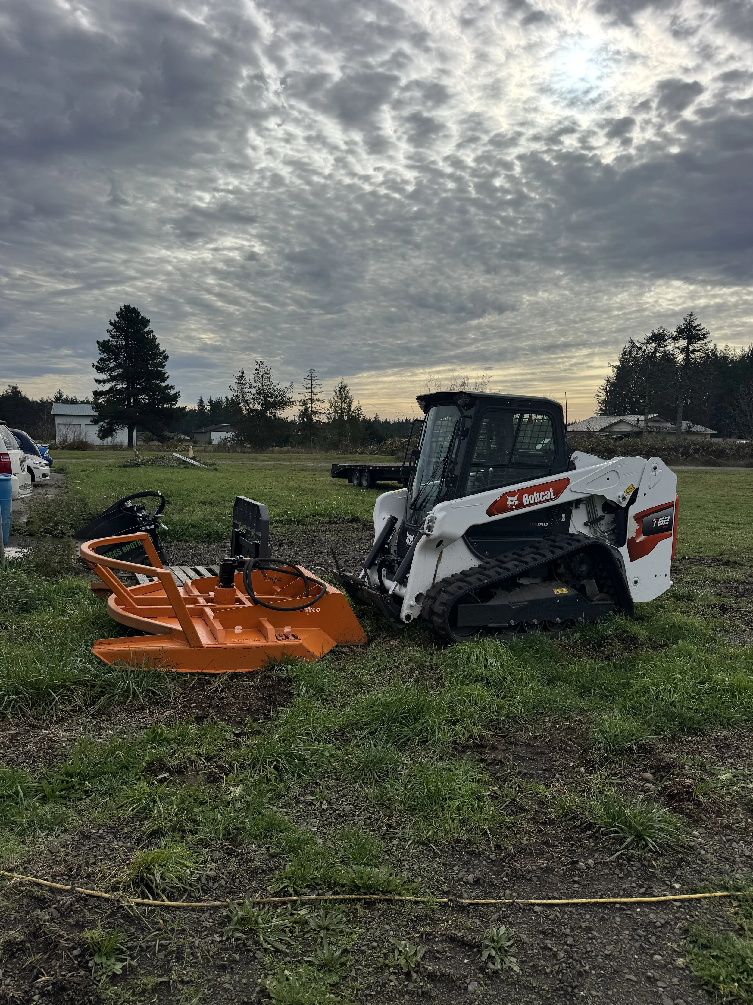 A Bobcat t62 track loader  with a Davco 72" brush hog on a grassy patch, under a cloudy sky with trees and buildings in the background.