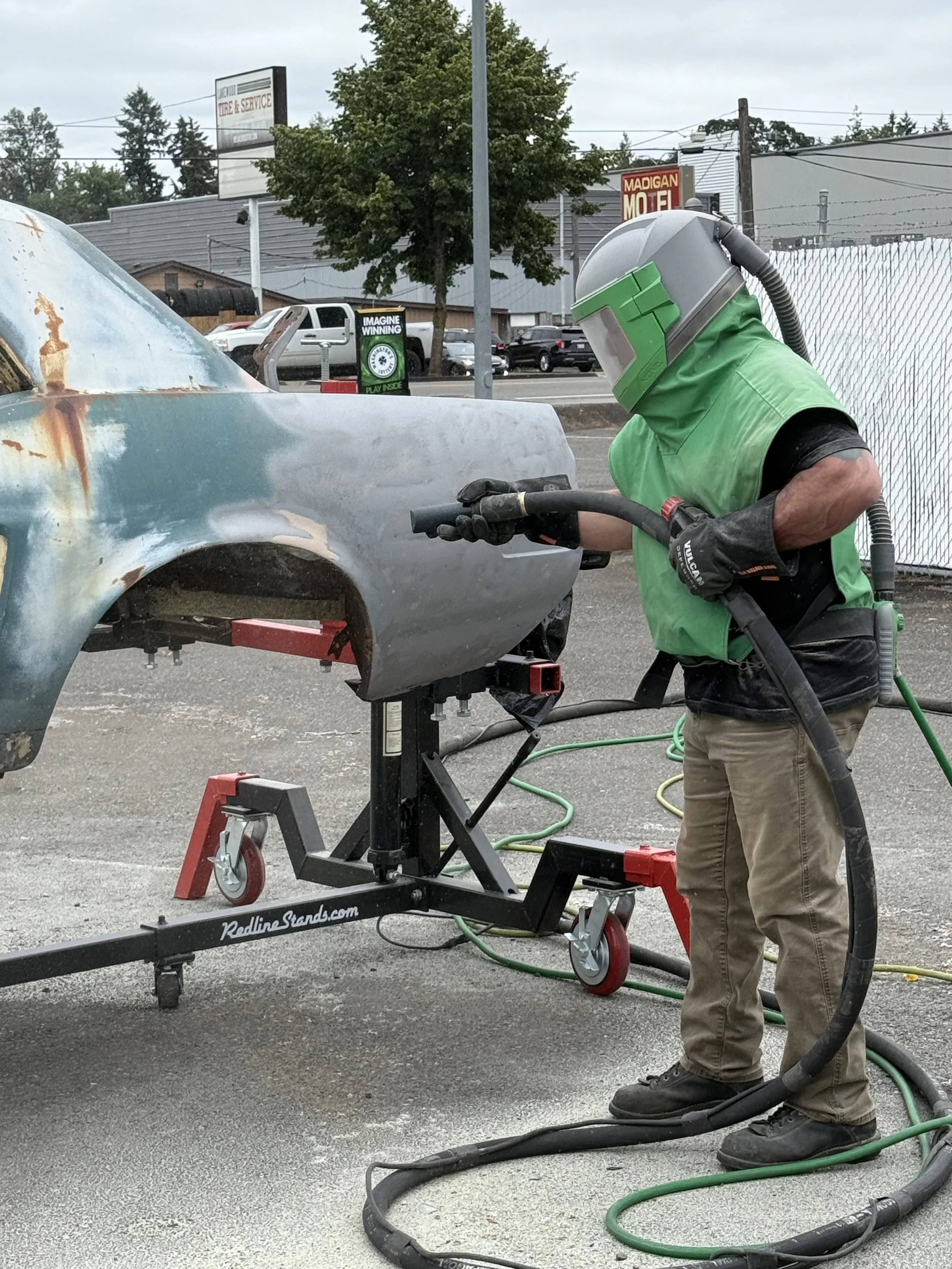 sand blasting a mustang, A person using a sandblaster to strip rust from an old car body, which is mounted on a stand in an outdoor lot.