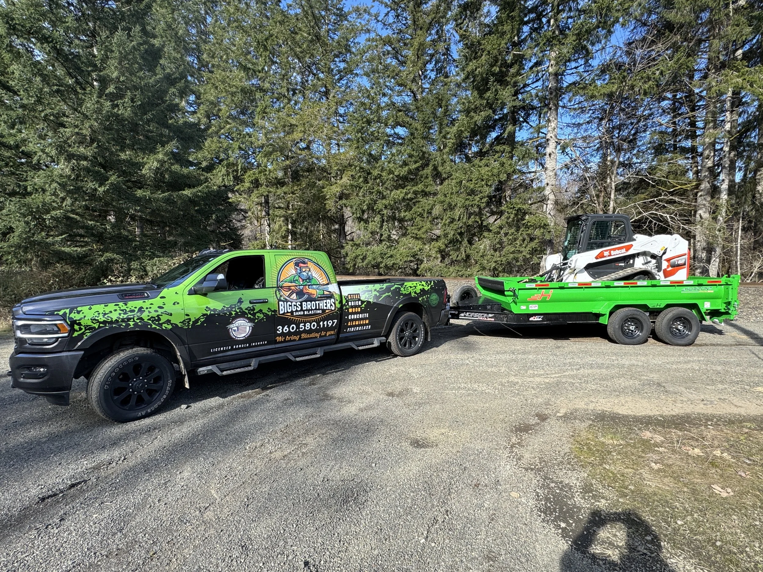 A black and green truck with advertising for Biggs Brothers Sand Blasting, pulling a trailer with a green flatbed, on which a white Bobcat skid-steer loader is loaded.