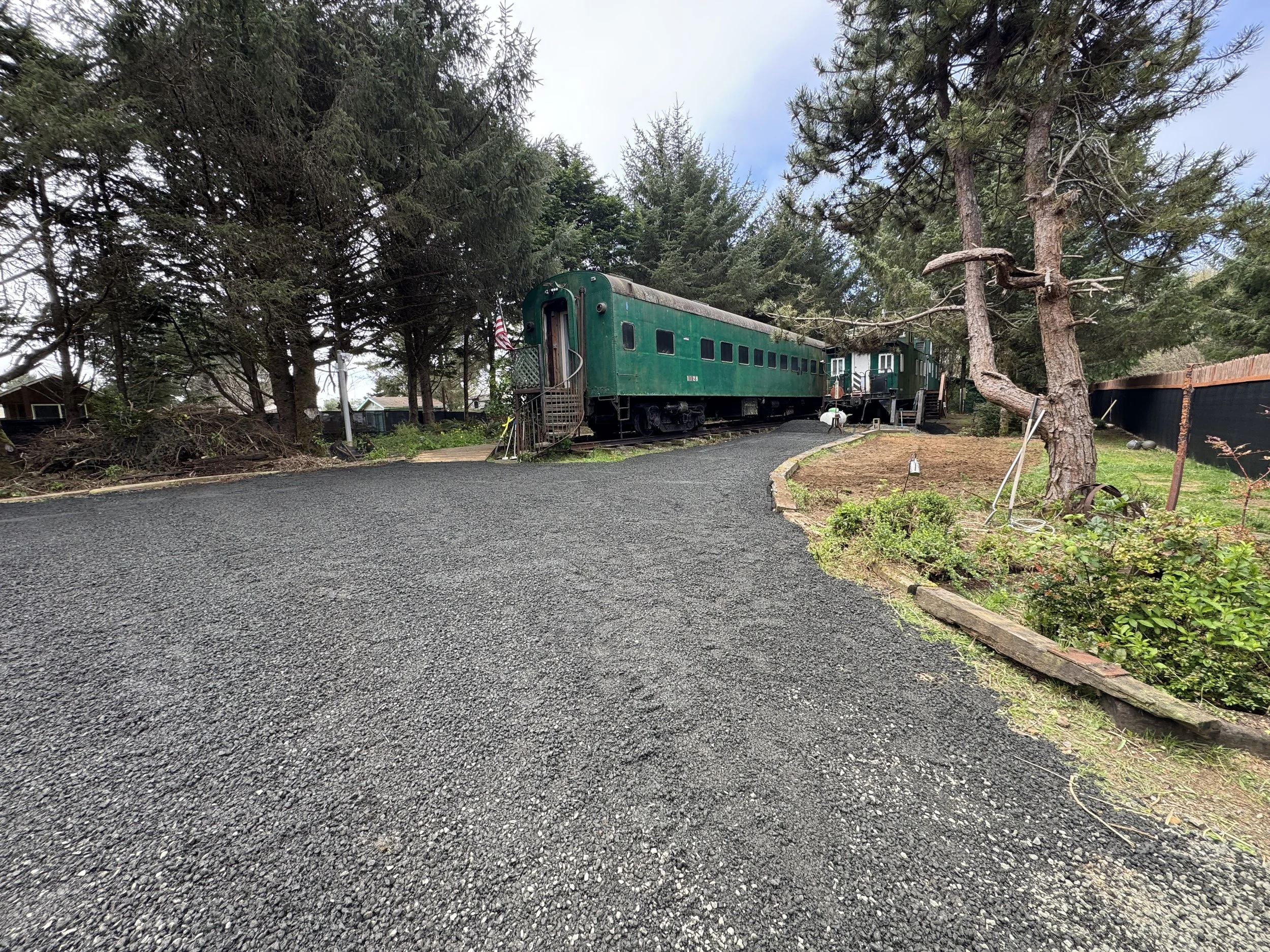A gravel driveway leading to a vintage green train car and a small train station nestled among tall pine trees and garden plants.