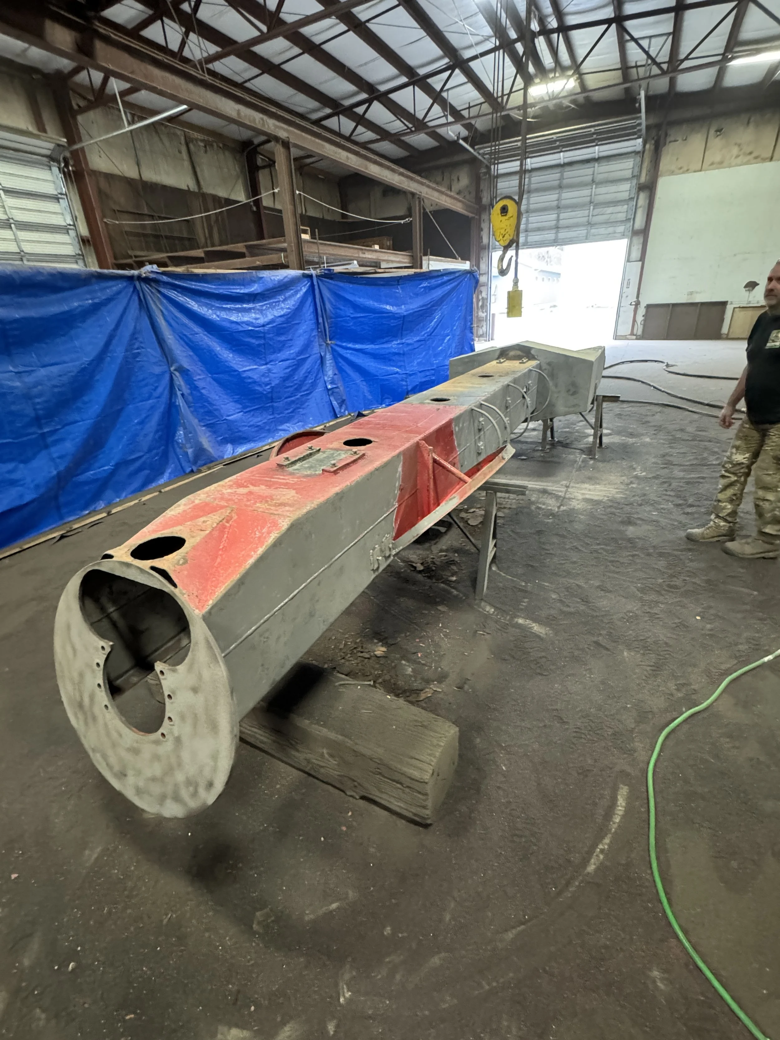 sand blasting An industrial workspace with a large cylindrical metal object on stands, some parts are painted red and gray, in a warehouse with a blue tarp and a person in camouflage pants and a black shirt nearby.