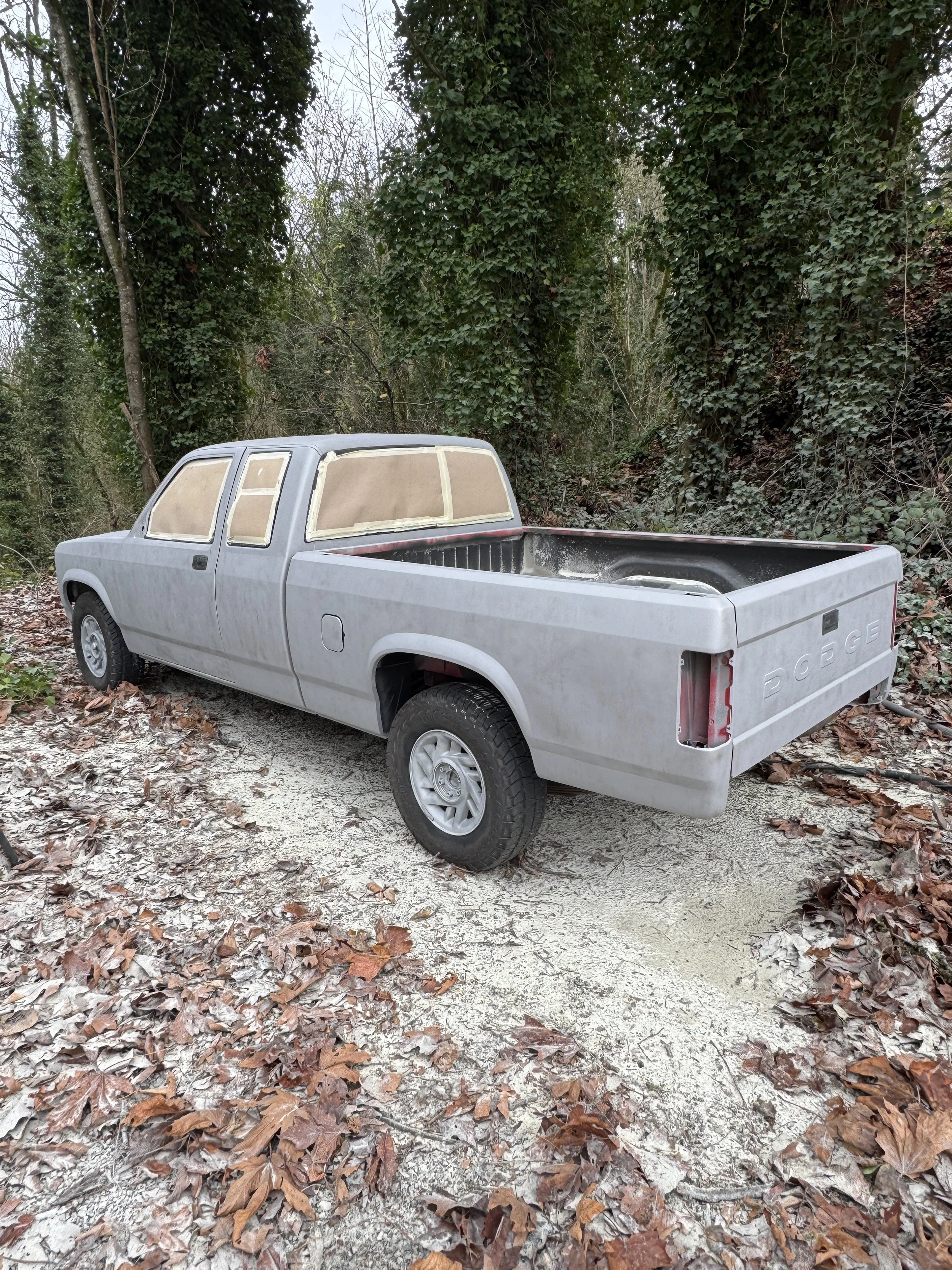 A Dodge Dakota pickup truck sand blasted outdoors, with its windows and body covered in a beige primer or paint, situated on a leaf-covered path in a wooded area.