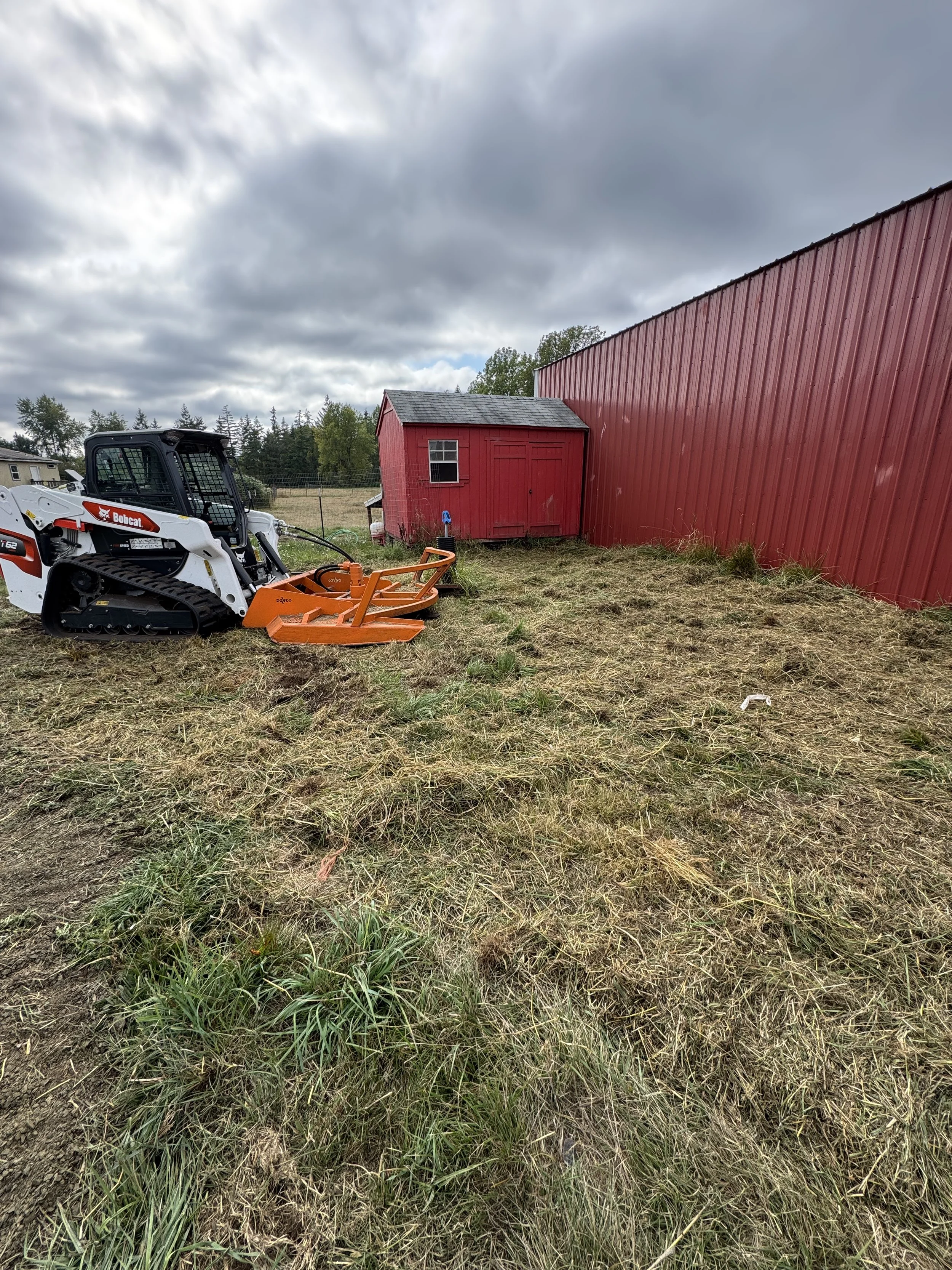 A small red shed and a larger red barn on a grassy lot under a cloudy sky, brushed hogged with a A Bobcat t62 track loader  with a Davco 72" brush hog parked nearby.