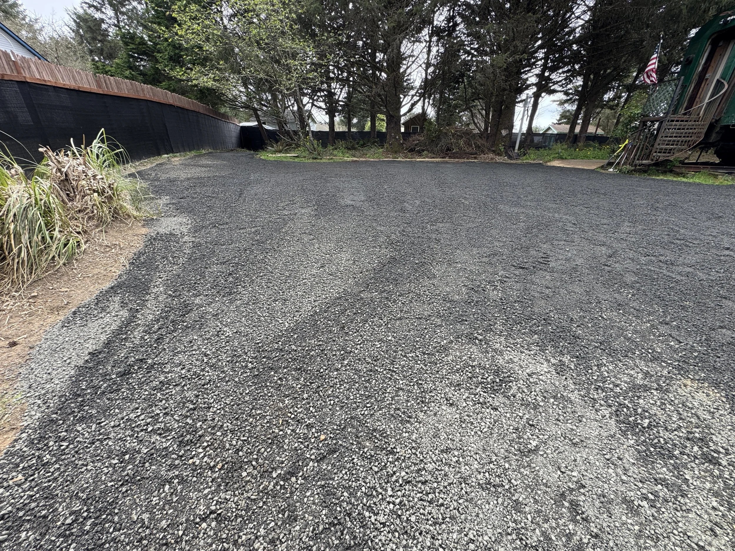 A gravel driveway with green plants on the left and trees in the background. There is a black fence on the left and a house on the right with an American flag and stairs.