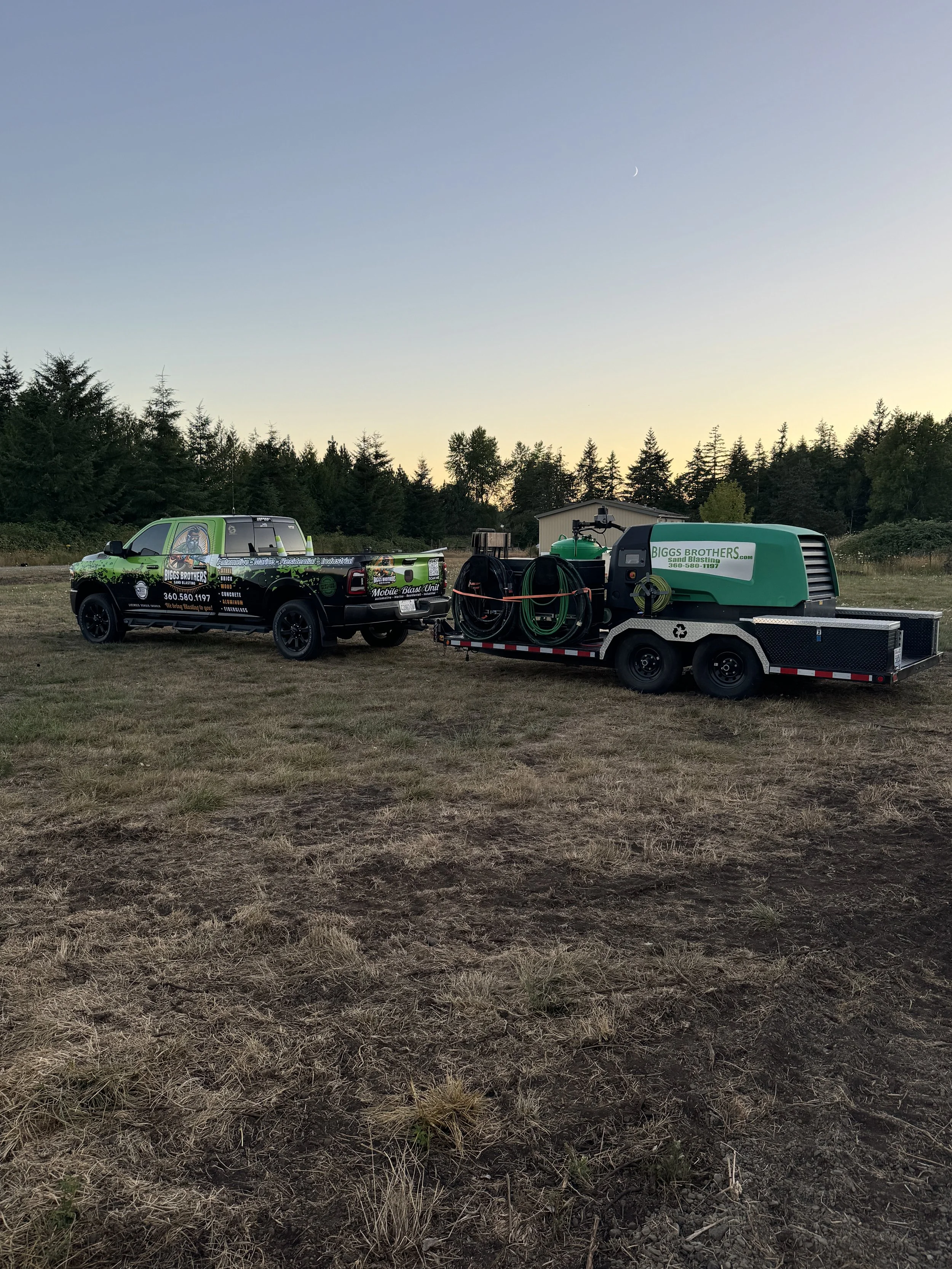 A black pickup truck with graphics and text advertising Biggs Brothers Sand Blasting, parked on a dirt field at dusk. The truck is attached to a trailer with a green sandblasting machine and hoses.