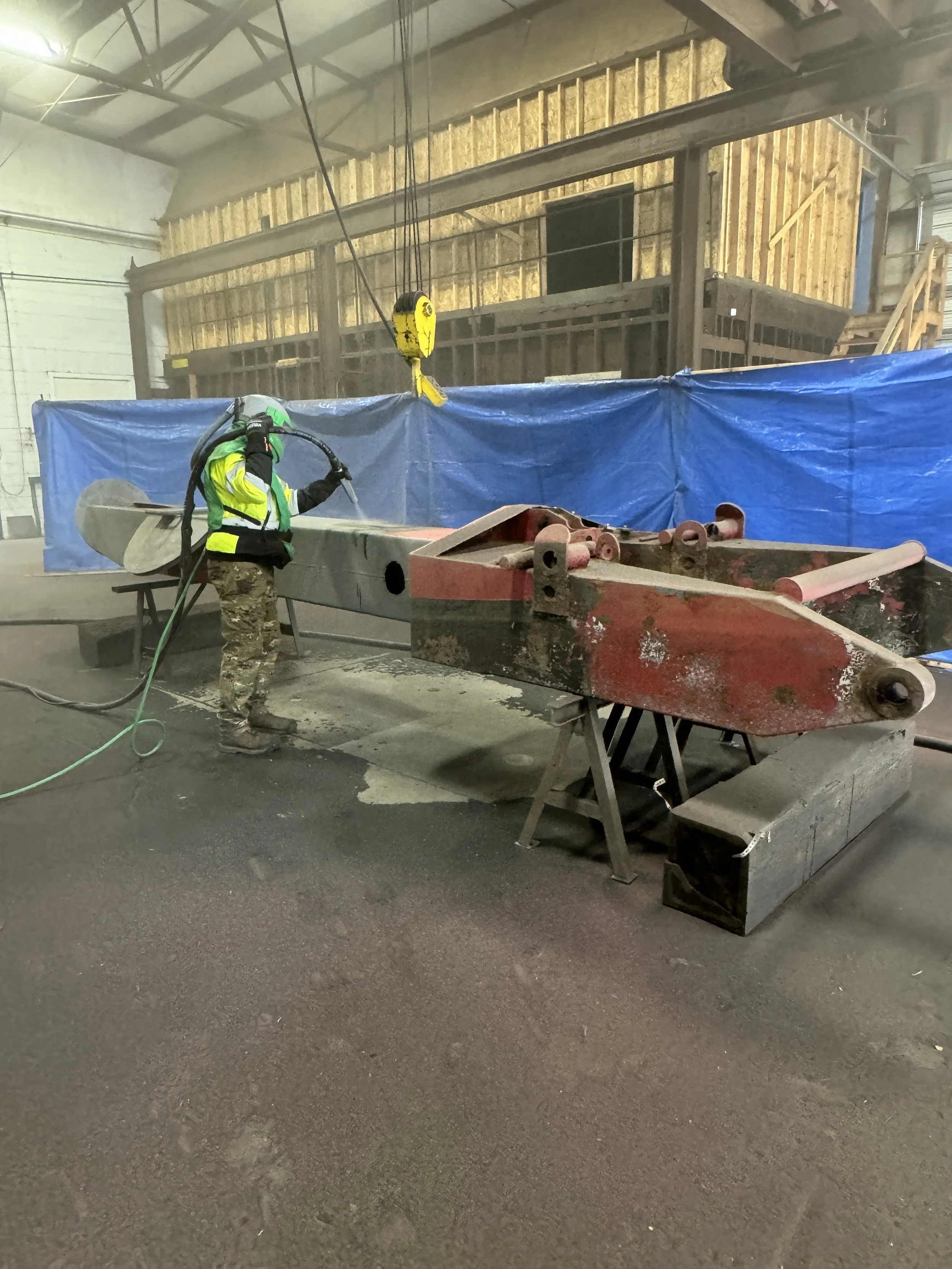 A worker in safety gear is sandblasting a large, metallic object inside an industrial workshop. The object is resting on supports, and there is a blue tarp in the background to contain debris.