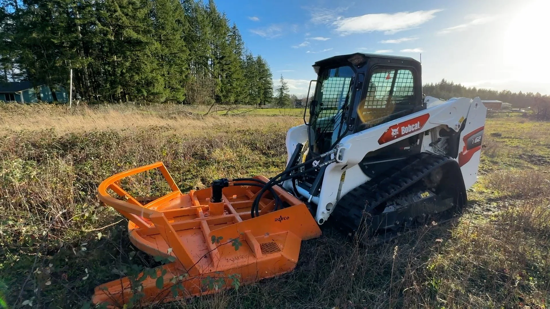 A Bobcat t62 compact track loader with an orange Davco Brush hog in a grassy field during daylight.