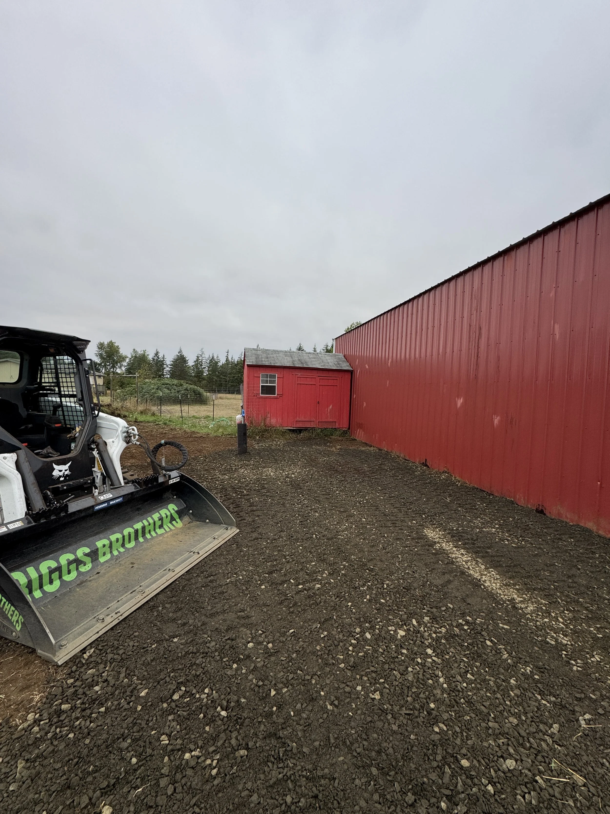 Red barn and small shed with a dirt and gravel yard, new grave graded, a t62 bobcat with a bucket attachment labeled quotes BIGGS BROTHERS