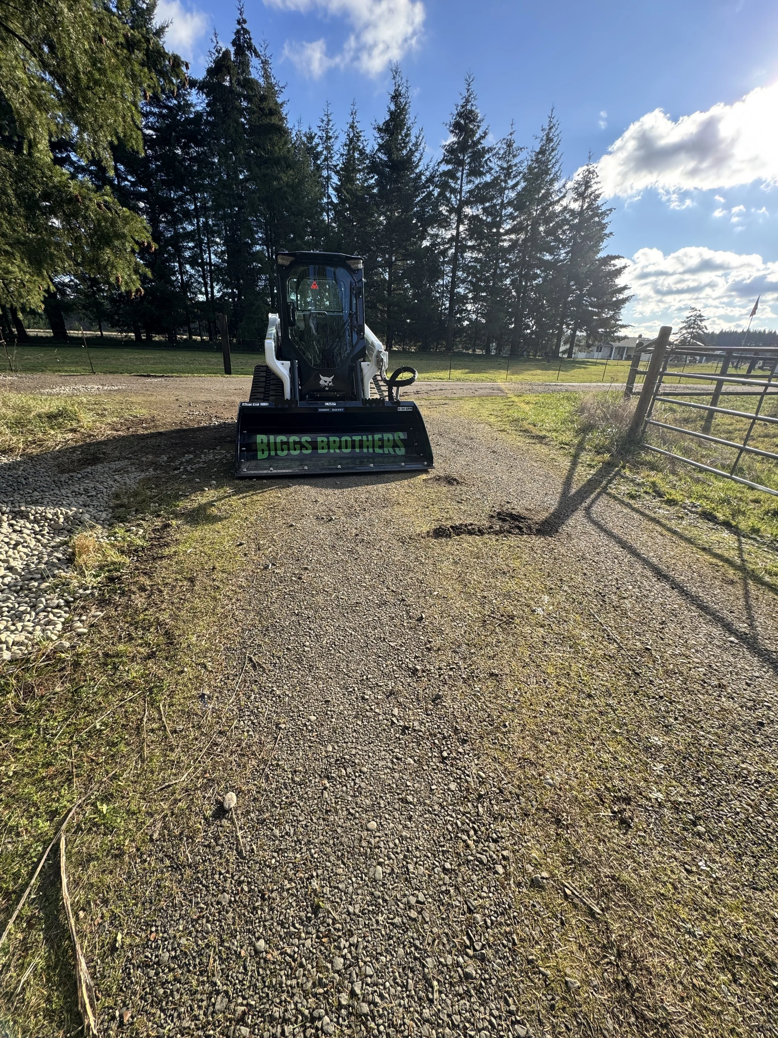 A Mini skid steer loader with a black blade attachment labeled 'BIGGS BROTHERS' parked on a gravel driveway surrounded by trees and a sunny sky.