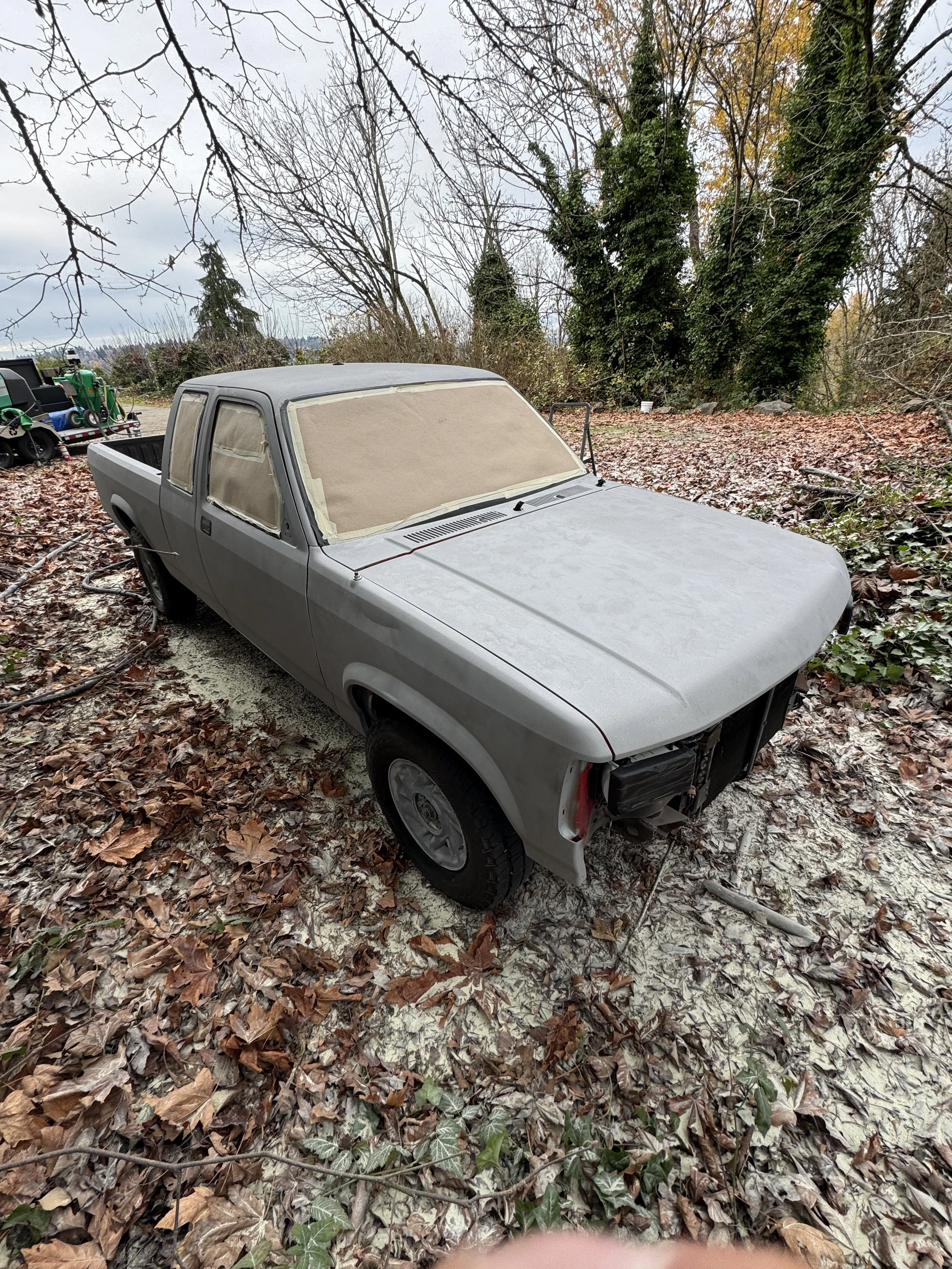A dodge dakota sandblasted pickup truck parked outdoors on a leaf-covered ground with no front grille, covered windows, and no front bumper, surrounded by trees with some leaves fallen and some still on the branches.