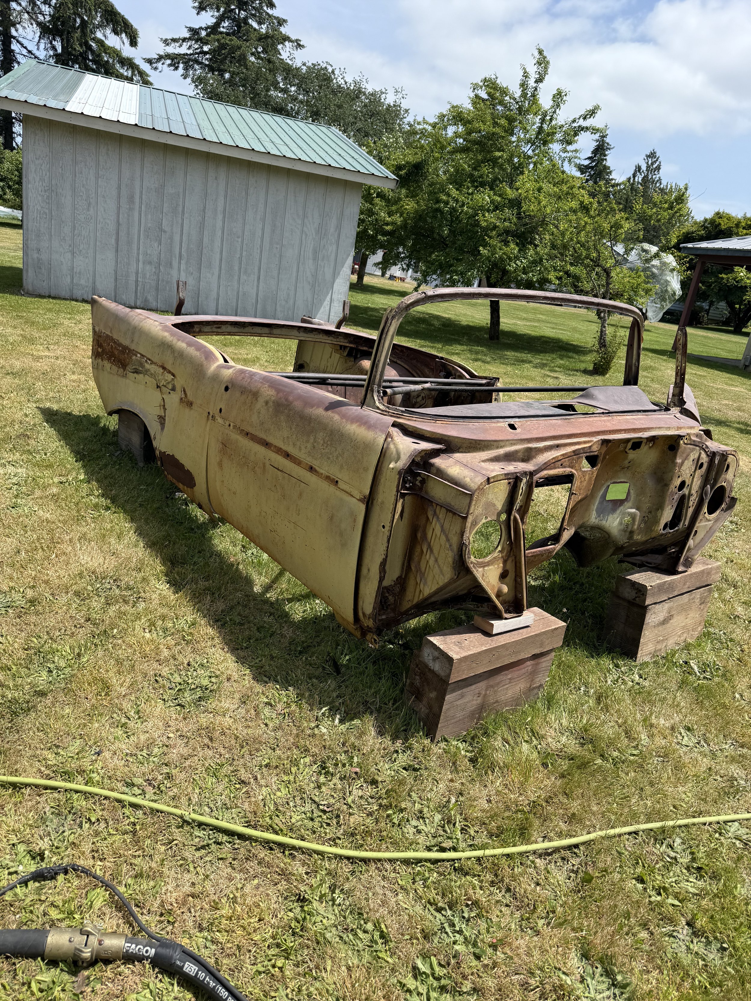 sand blasted a 57 chevy, Rusty, vintage car body resting on wooden blocks in a grassy yard with trees and a shed in the background.