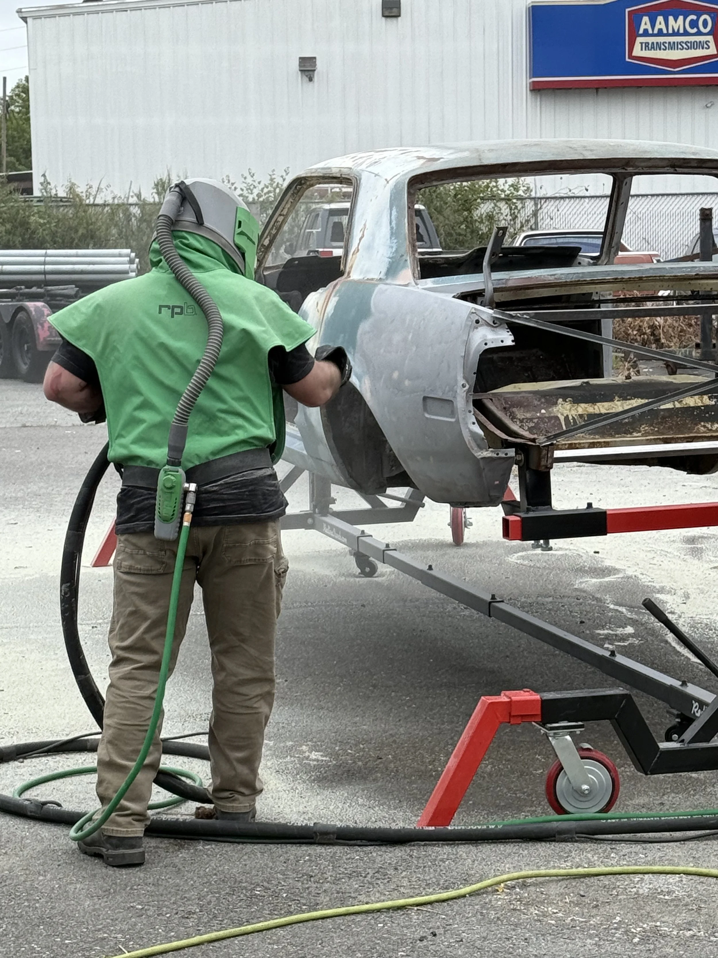 Sand blasting a 65 mustang, A worker wearing a green respirator and shirt cleaning or working on an old, rusted car body outside near a building with an AMCO Transmissions sign, using a vacuum or spray equipment.