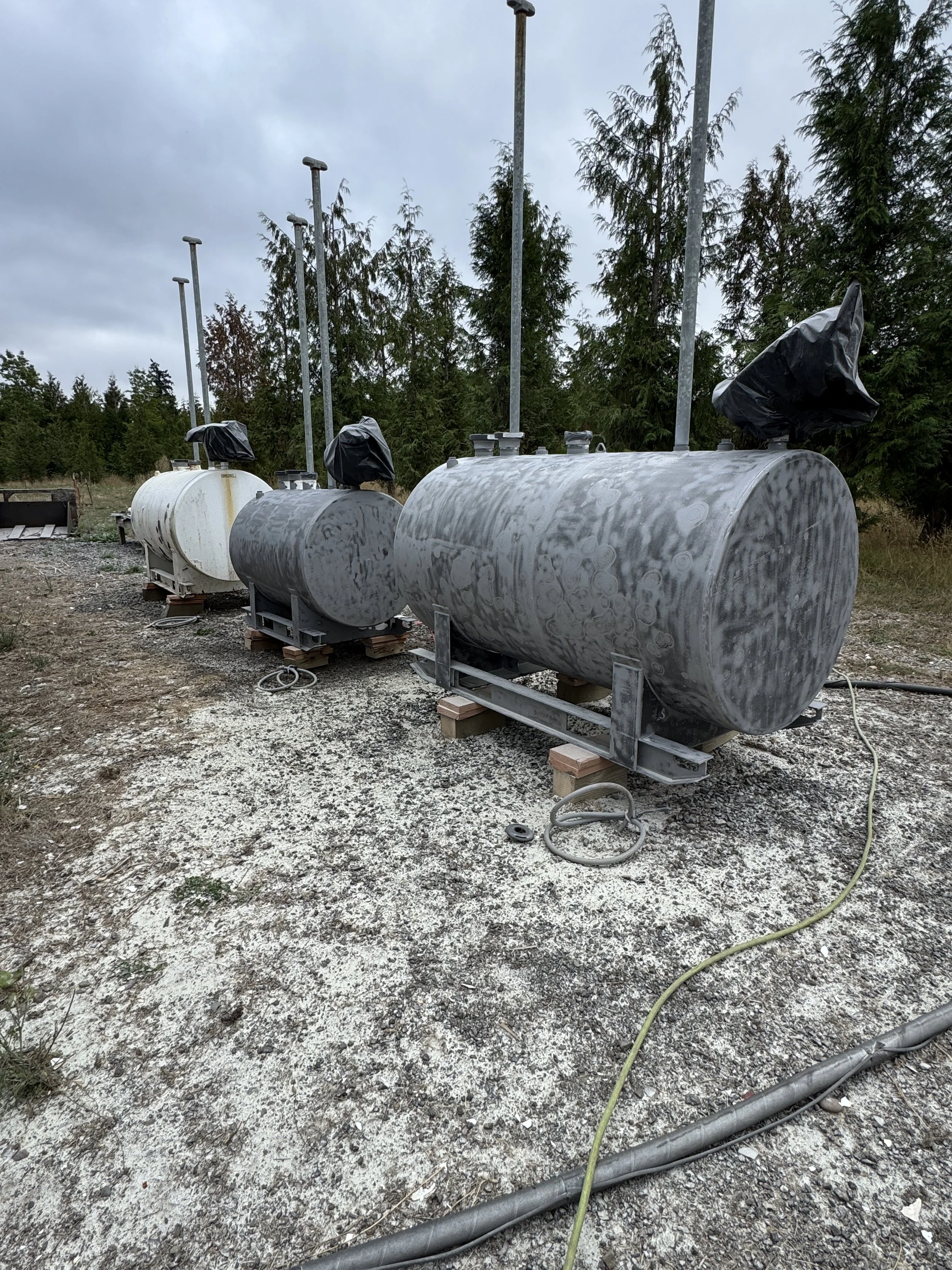 Sand blasted Three large industrial storage tanks on concrete pads outdoors, with a wooded background and cloudy sky.