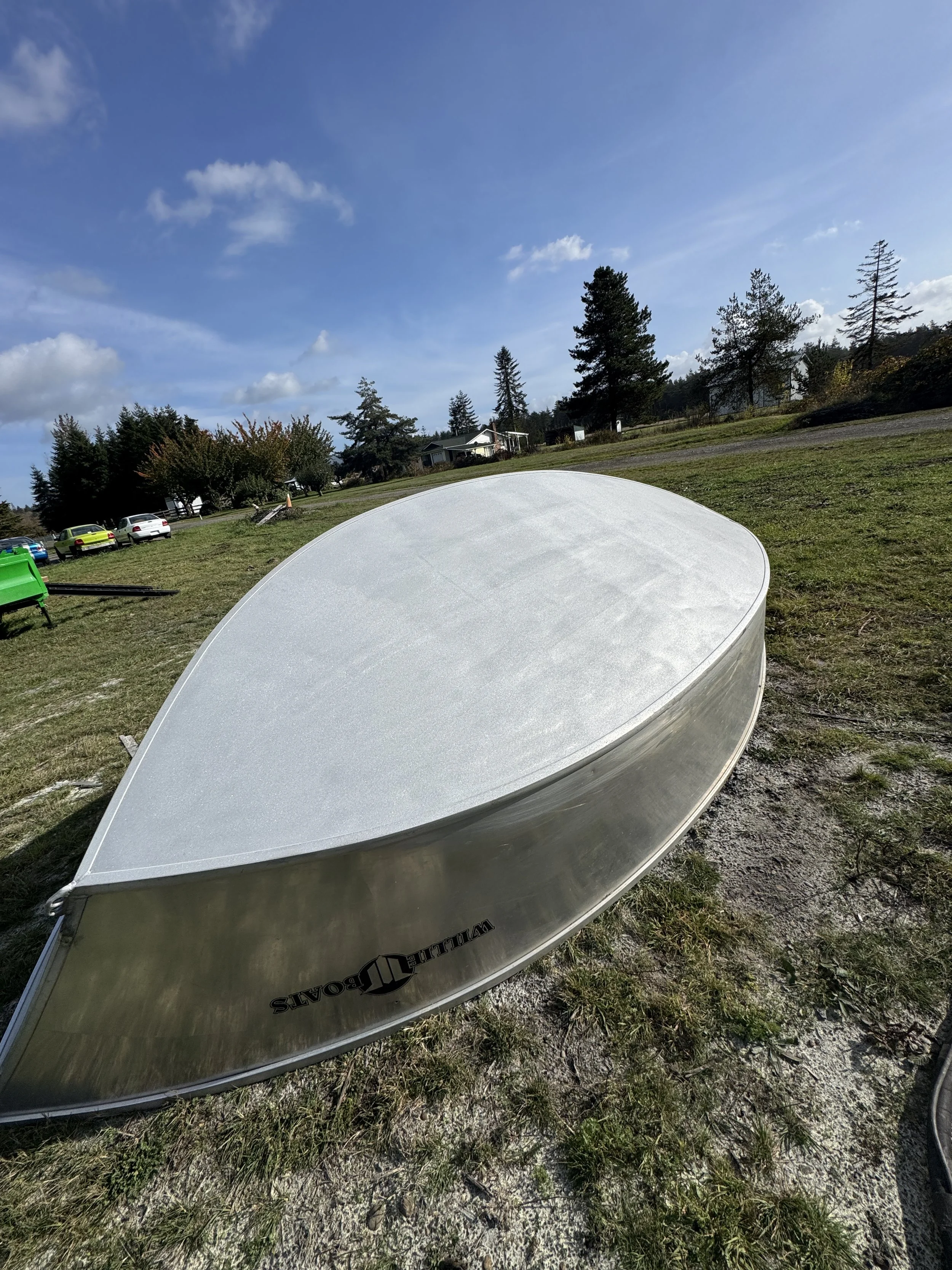 Upturned sand blasted boat bottom lying on grass and dirt, with trees, houses, and cars in the background under a partly cloudy blue sky.