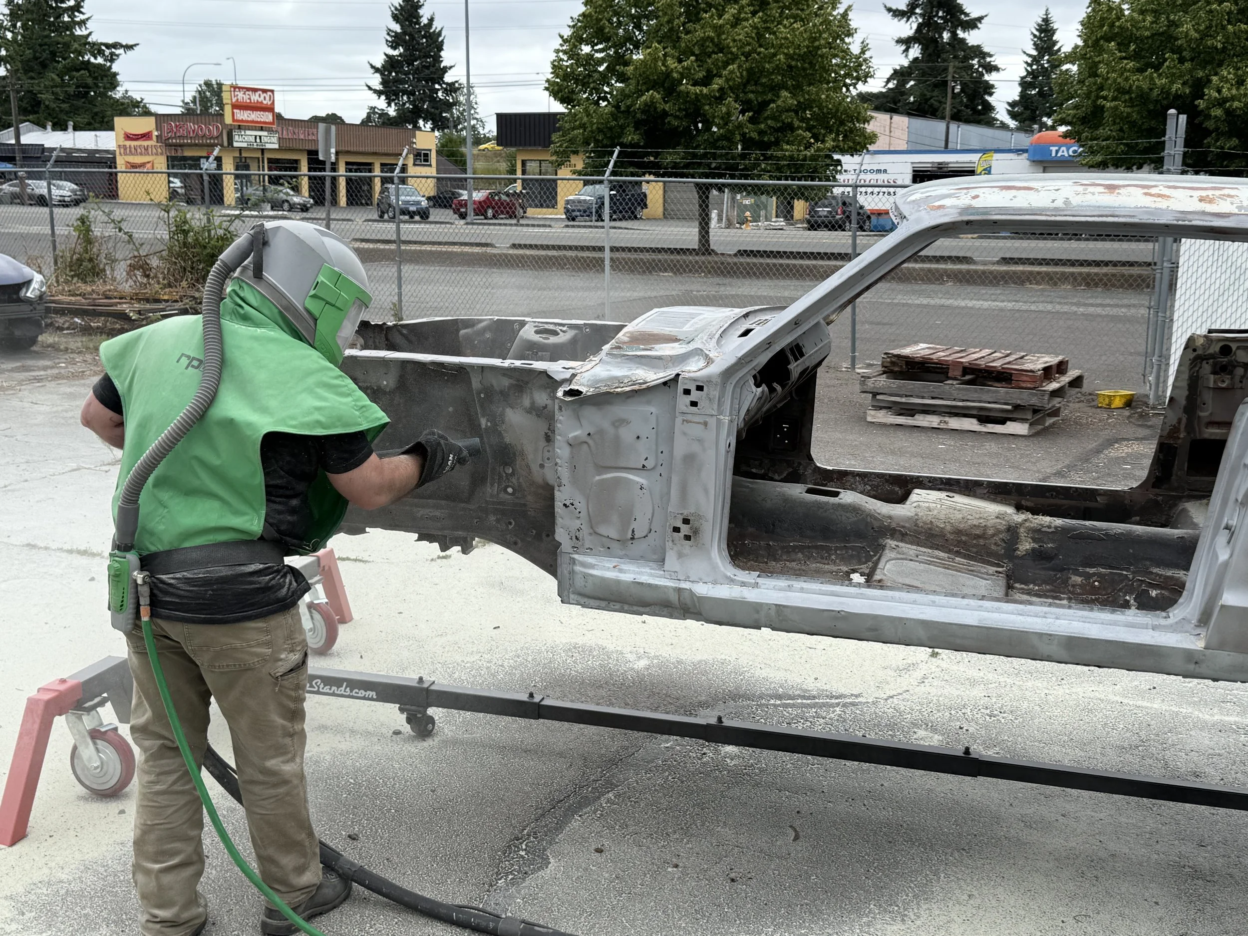 Sand blasting a mustang A worker in safety gear, including a helmet and gloves, is using a tool on a metallic vehicle frame outside, with a chain-link fence and commercial buildings in the background.
