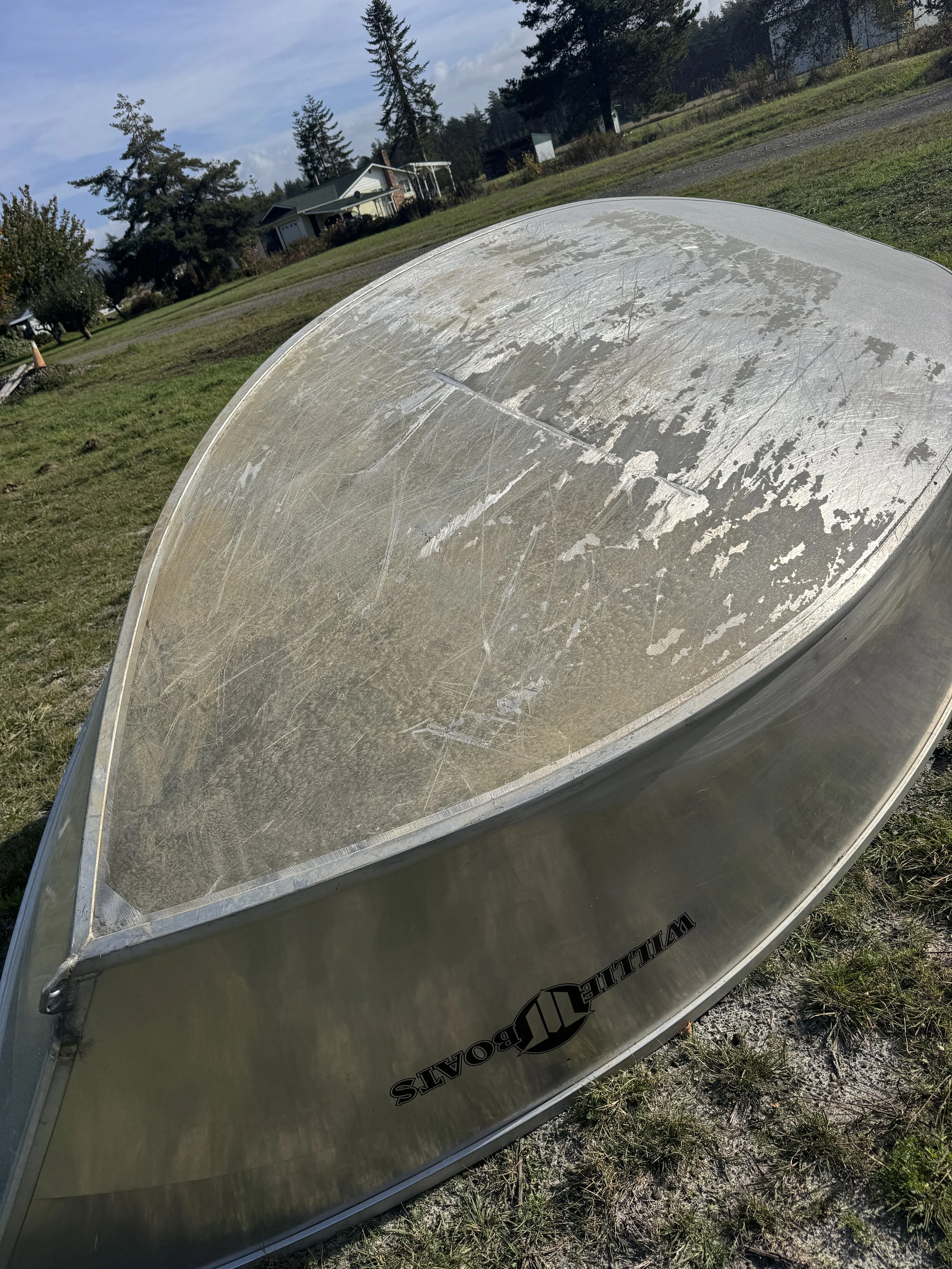 A weathered aluminum boat with a scratched and dirty surface partially sand blasted, resting on grass outdoors in a rural area.