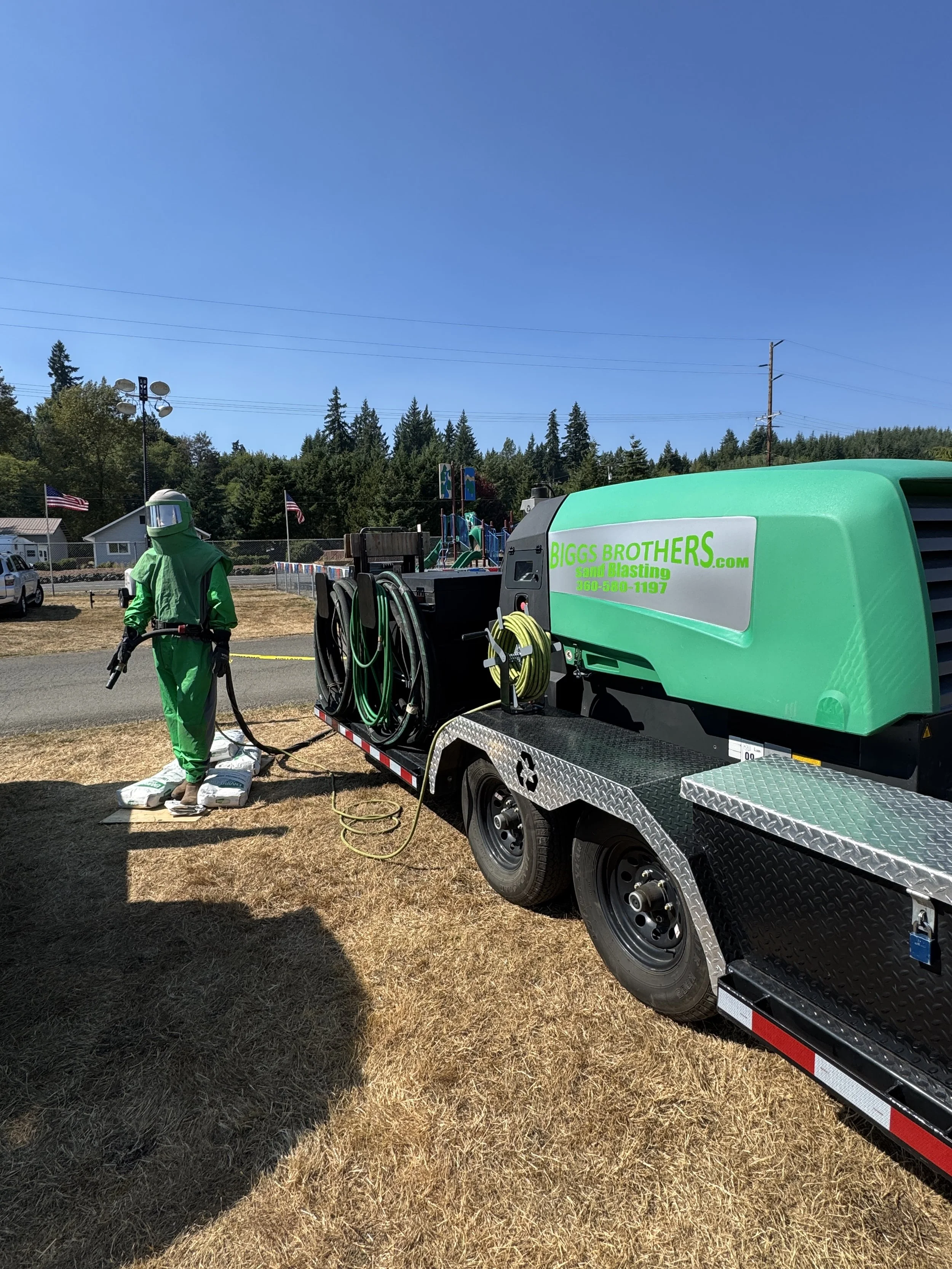Person in green sand blasting gear and mask near a truck labeled 'BIGGS BROTHERS' on a grassy area with a playground and American flags in the background.