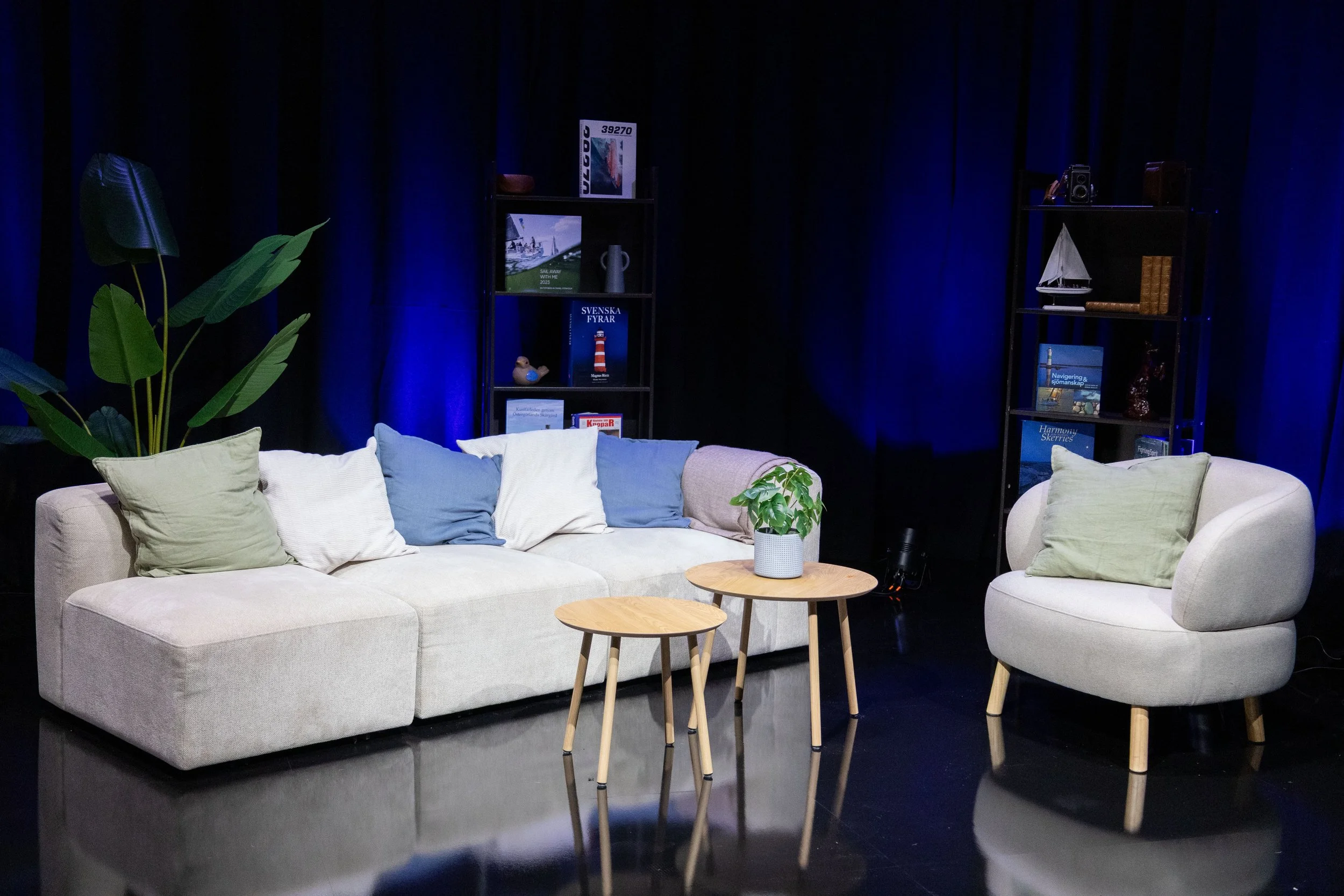 A living room with a light beige sofa and an armchair, both with green and blue cushions, two small round garden tables with a plant on one, black theater or studio lighting, dark background with books and decorations on shelves.