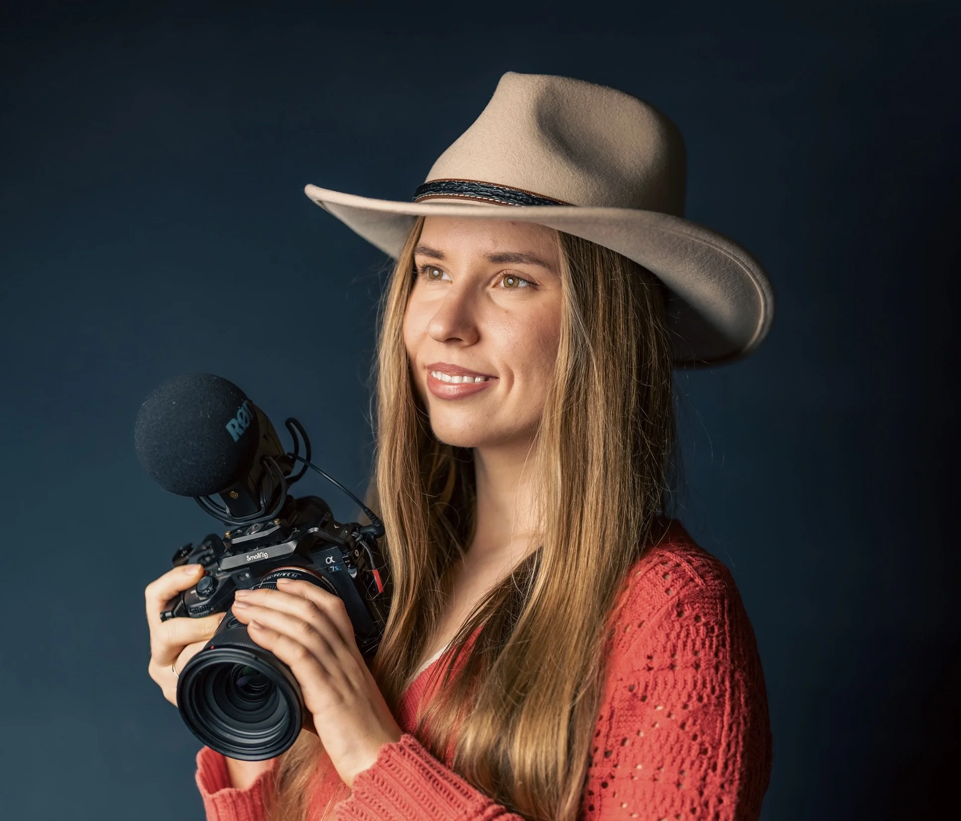 A woman wearing a beige cowboy hat and a red sweater holding a camera with a microphone attached, against a dark background.