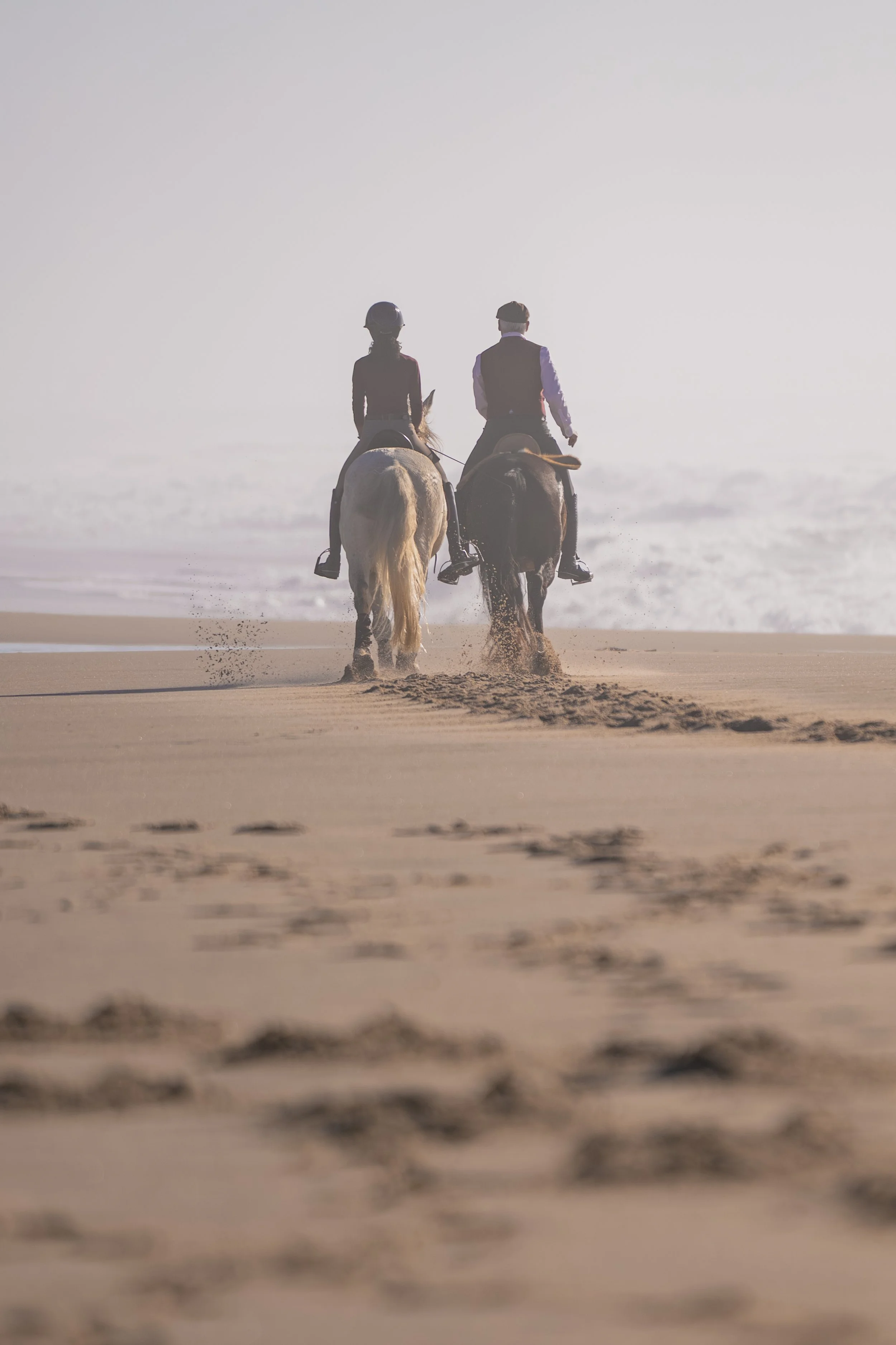 Two people riding lusitano horses along the beach in Portugal at sunset or sunrise, leaving footprints in the sand. This photo was made by Maria Steppe from Wonderveld Pictures.