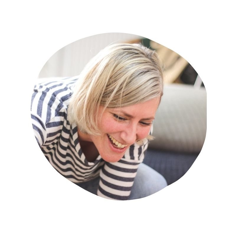 Lachende Frau mit blonden Haaren und gestreiftem Pullover, sitzend auf einem Sofa.